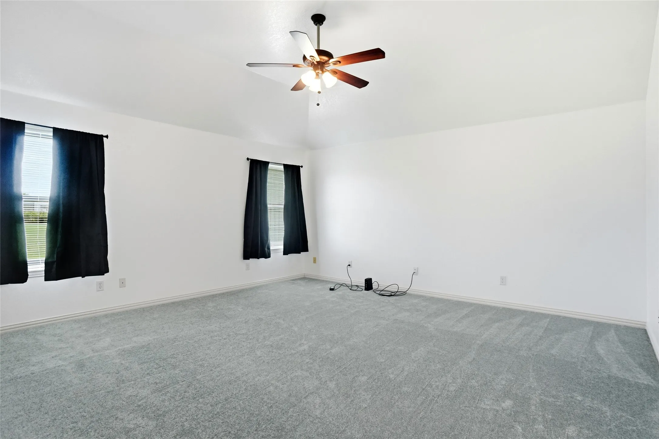 Spacious room featuring vaulted ceilings, light gray carpeting, two windows with blinds, and a ceiling fan with integrated lighting