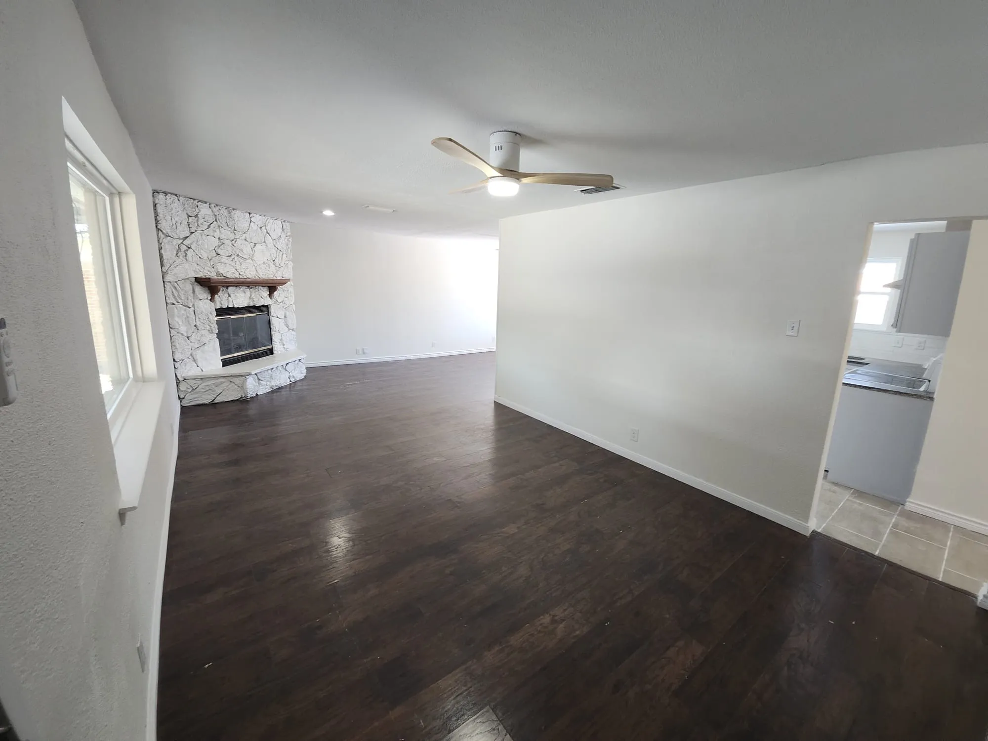 Spacious interior featuring dark wood-finish flooring, a white stone fireplace with a wood mantel, and a ceiling fan with integrated lighting