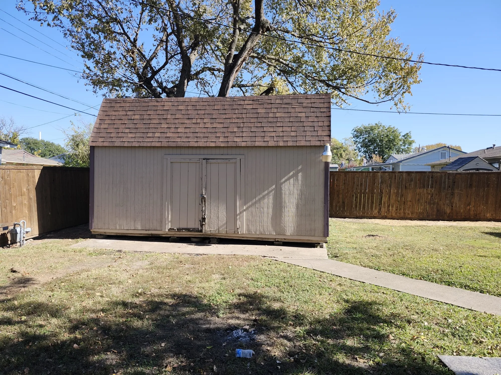 Gable-roof storage shed with double doors situated on a concrete pad, surrounded by a wood privacy fence and a grassy area with a partial concrete path