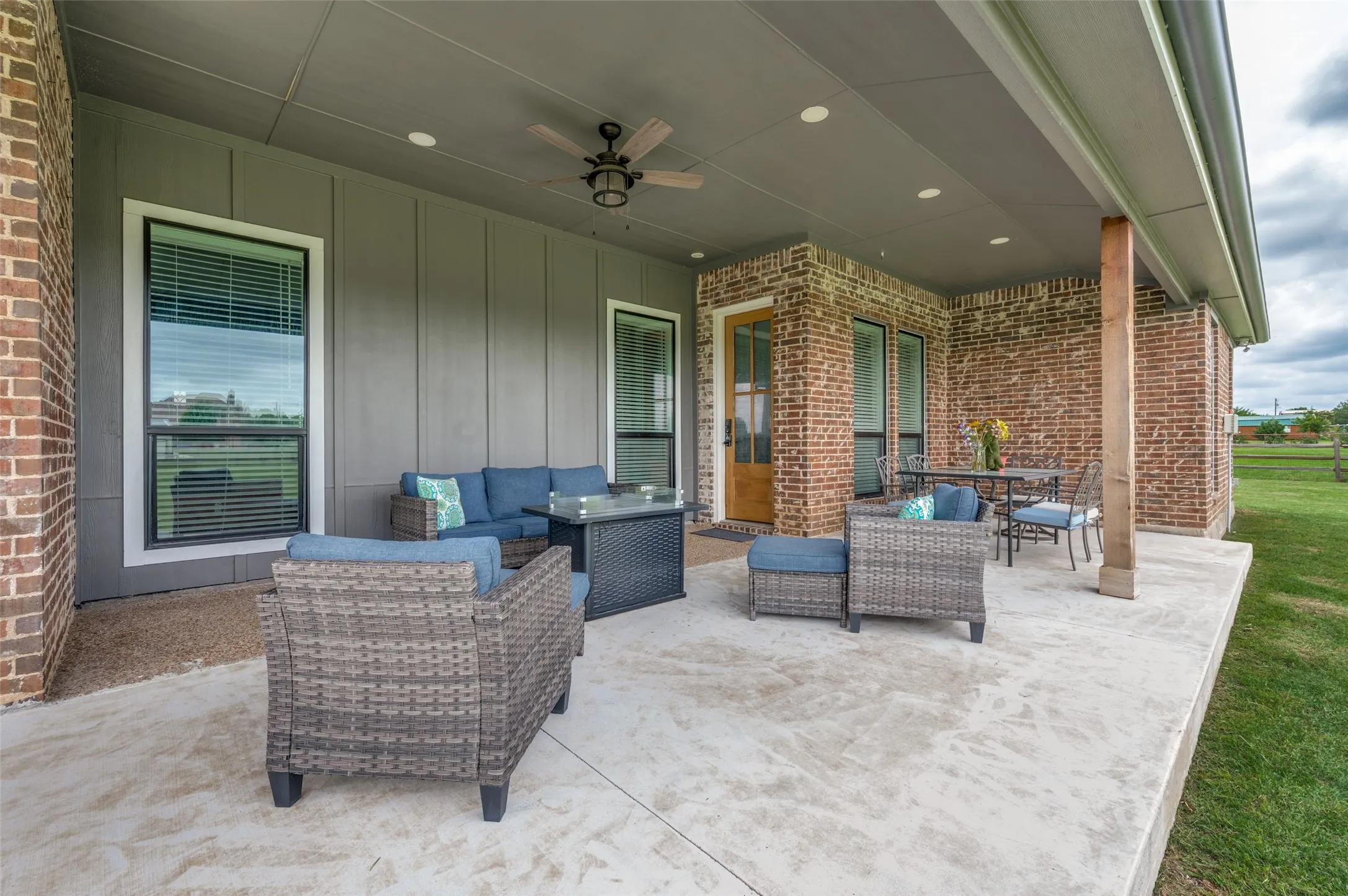 Outdoor living space at this expansive covered patio, thoughtfully designed for year-round enjoyment. With ample room for both lounge seating and outdoor dining, this inviting retreat is perfect for hosting gatherings, enjoying family meals, or simply relaxing while taking in the peaceful surroundings. The soaring covered ceiling features recessed lighting and a ceiling fan for added comfort, while the combination of brick and board-and-batten accents creates a warm, upscale atmosphere. Large windows and direct access to the home's main living areas make indoor-outdoor entertaining effortless. Whether sipping your morning coffee, grilling with friends, or unwinding at sunset, this spacious outdoor living area offers the perfect blend of comfort, style, and functionality.