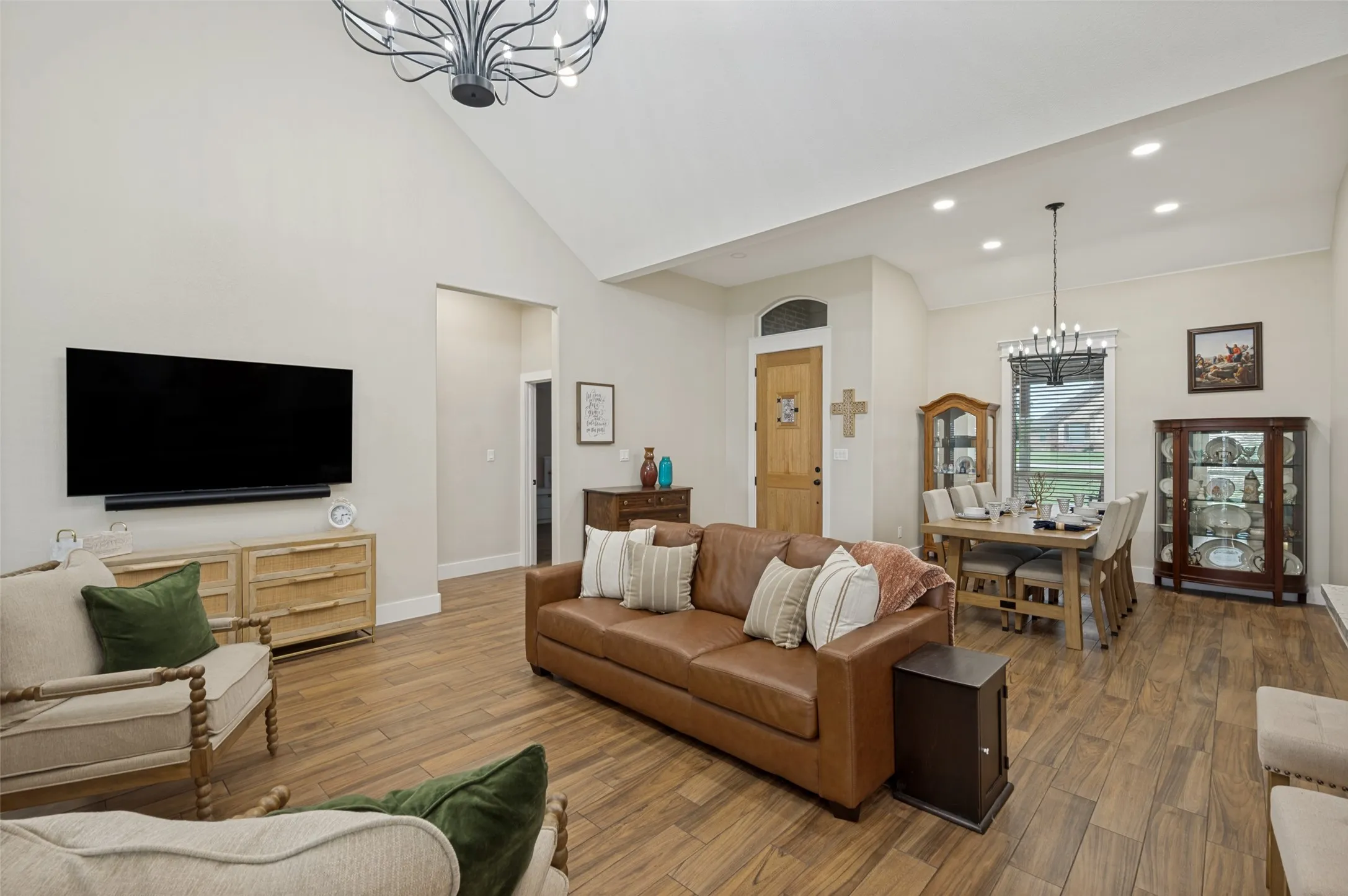 As you step inside, you're welcomed by an inviting open-concept living space highlighted by soaring vaulted ceilings, rich wood-look flooring, and abundant natural light. The spacious living room offers a warm yet modern feel, with clean lines, neutral tones, and elegant lighting that create an airy atmosphere. Beautiful formal dining room has a scaled-down chandelier that matches the larger one in the family room. The open area to the left in this photo leads to the guest wing.  Notice the beautiful entry door with transom window above.