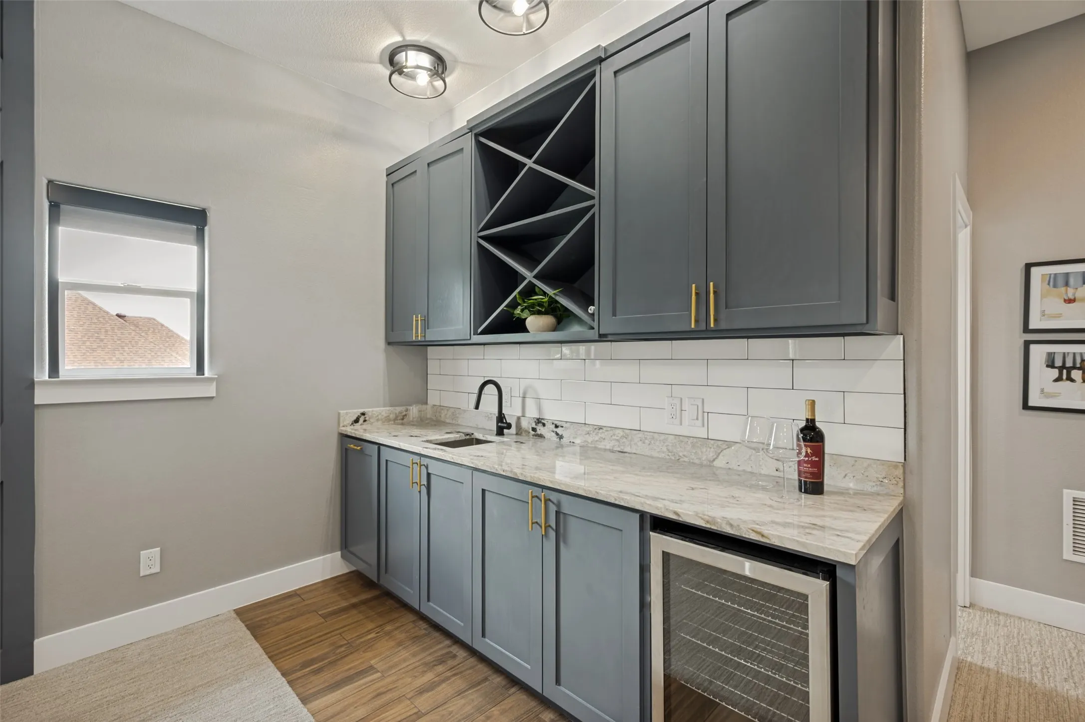 Perfect for entertaining, this stylish upstairs wet bar adds both convenience and sophistication to the bonus room. Custom cabinetry in a rich designer finish is paired with elegant granite countertops, creating a polished and functional space for serving guests. The built-in beverage refrigerator keeps drinks perfectly chilled, while the sink and ample counter space make hosting effortless. A striking geometric wine rack provides both storage and visual appeal, while the subway tile backsplash and brushed gold hardware add a touch of modern elegance. Wine refrigerator stays.