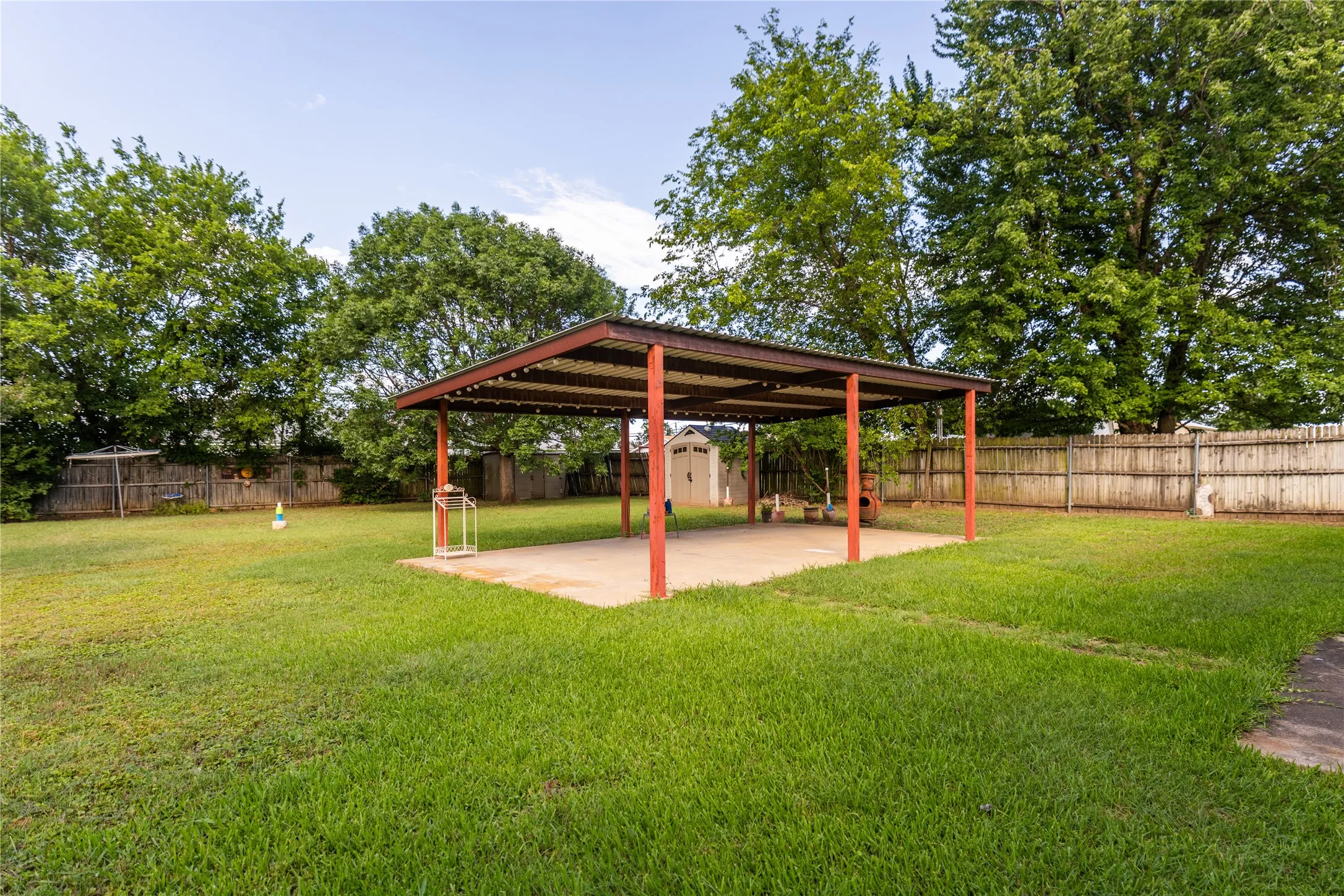 Expansive backyard featuring a covered patio structure with a concrete base, a detached storage shed, a wood privacy fence, and mature trees lining the perimeter