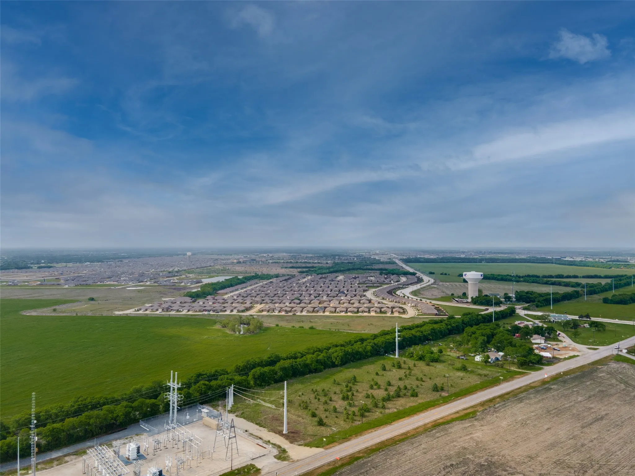 Expansive aerial perspective showcasing a developed residential area, undeveloped green fields, a water tower, and a power substation
