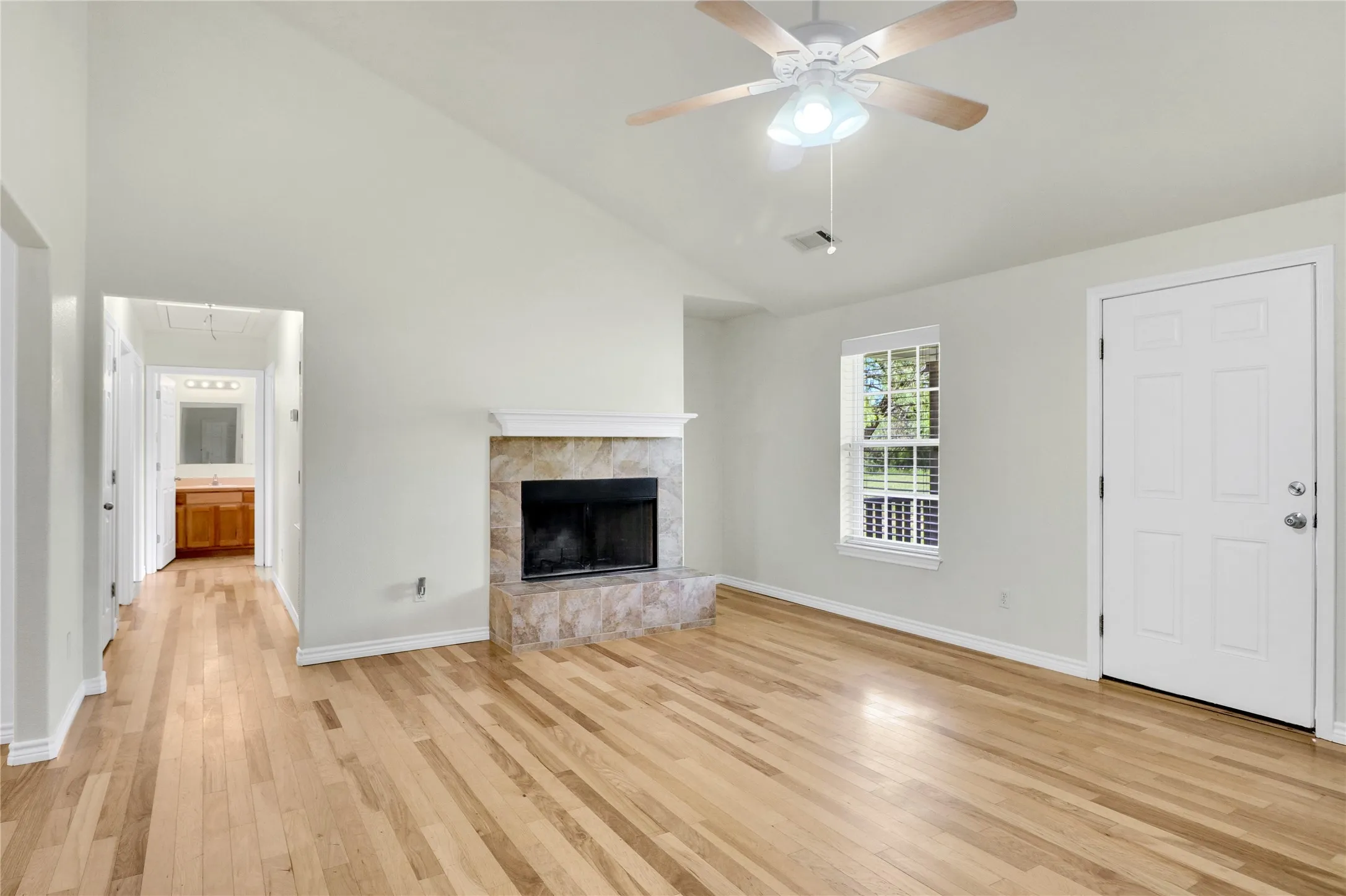Vaulted ceiling, wood-finish flooring, stone-faced fireplace with white mantel, ceiling fan with light fixture, and window with blinds