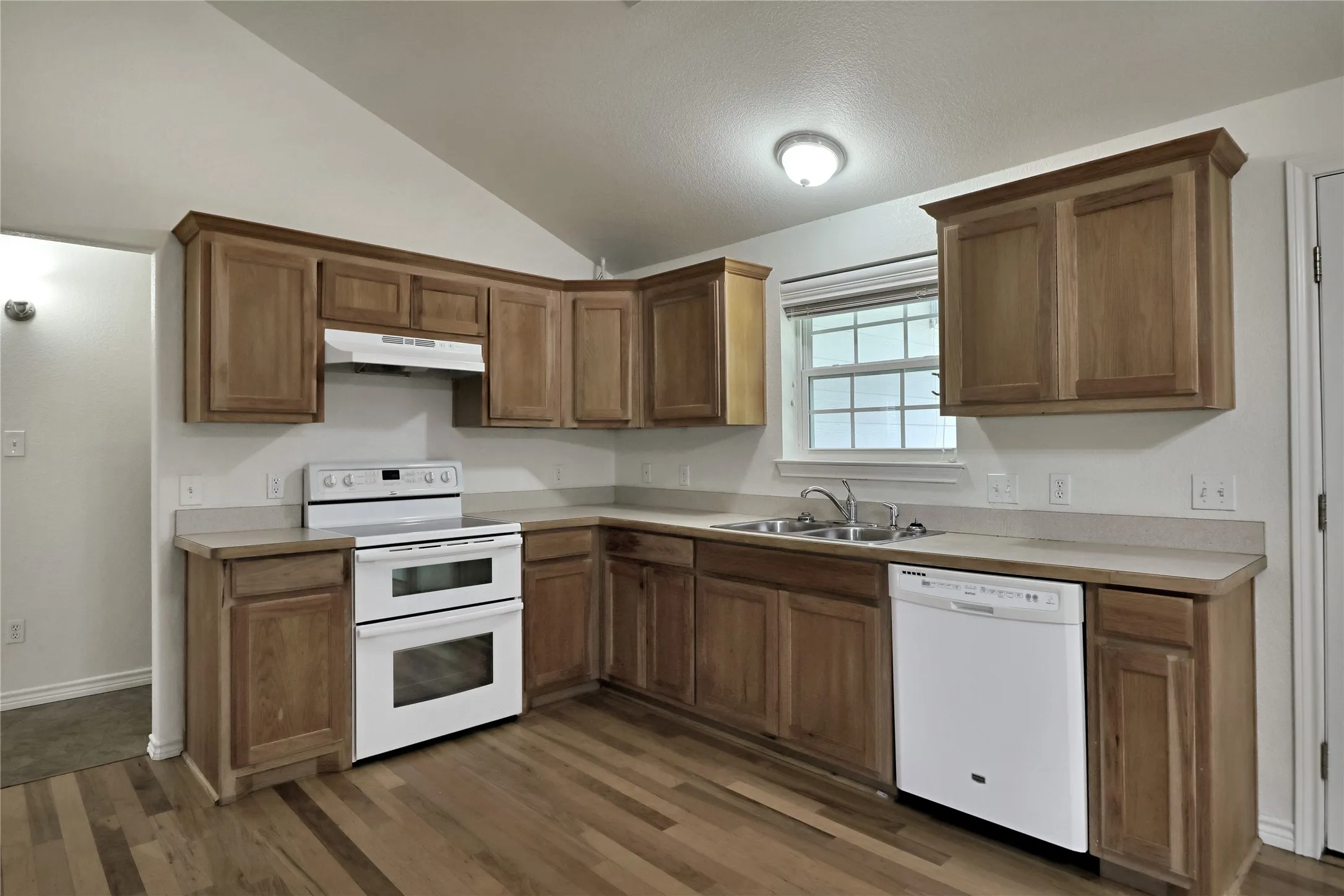 Kitchen featuring wood-finish flooring, L-shaped cabinetry, light-toned countertops, double oven range, and a double basin sink beneath a window