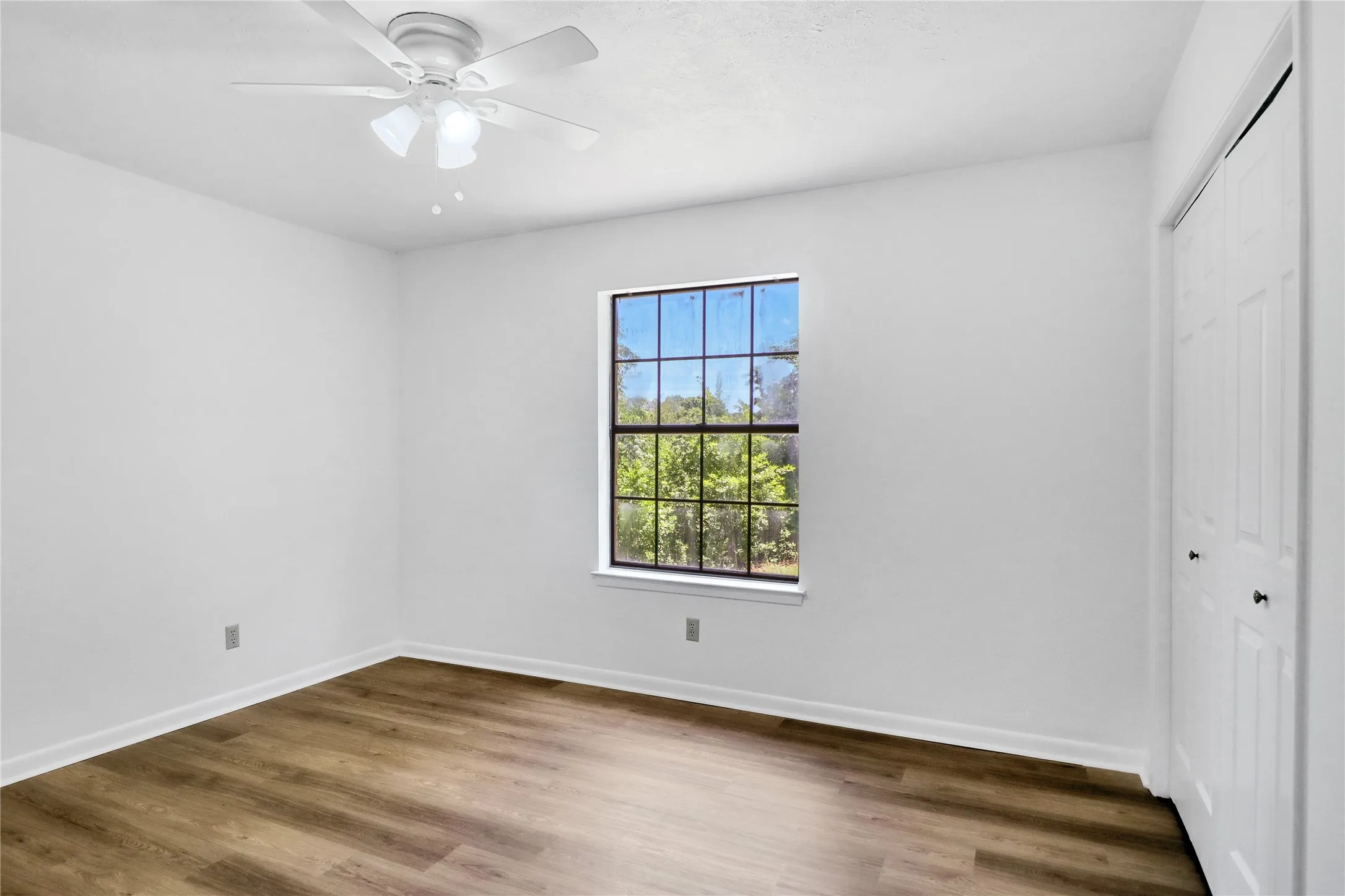 Spacious room featuring wood-finish flooring, a ceiling fan with integrated lighting, a window with exterior visibility, and a built-in closet with bi-fold doors