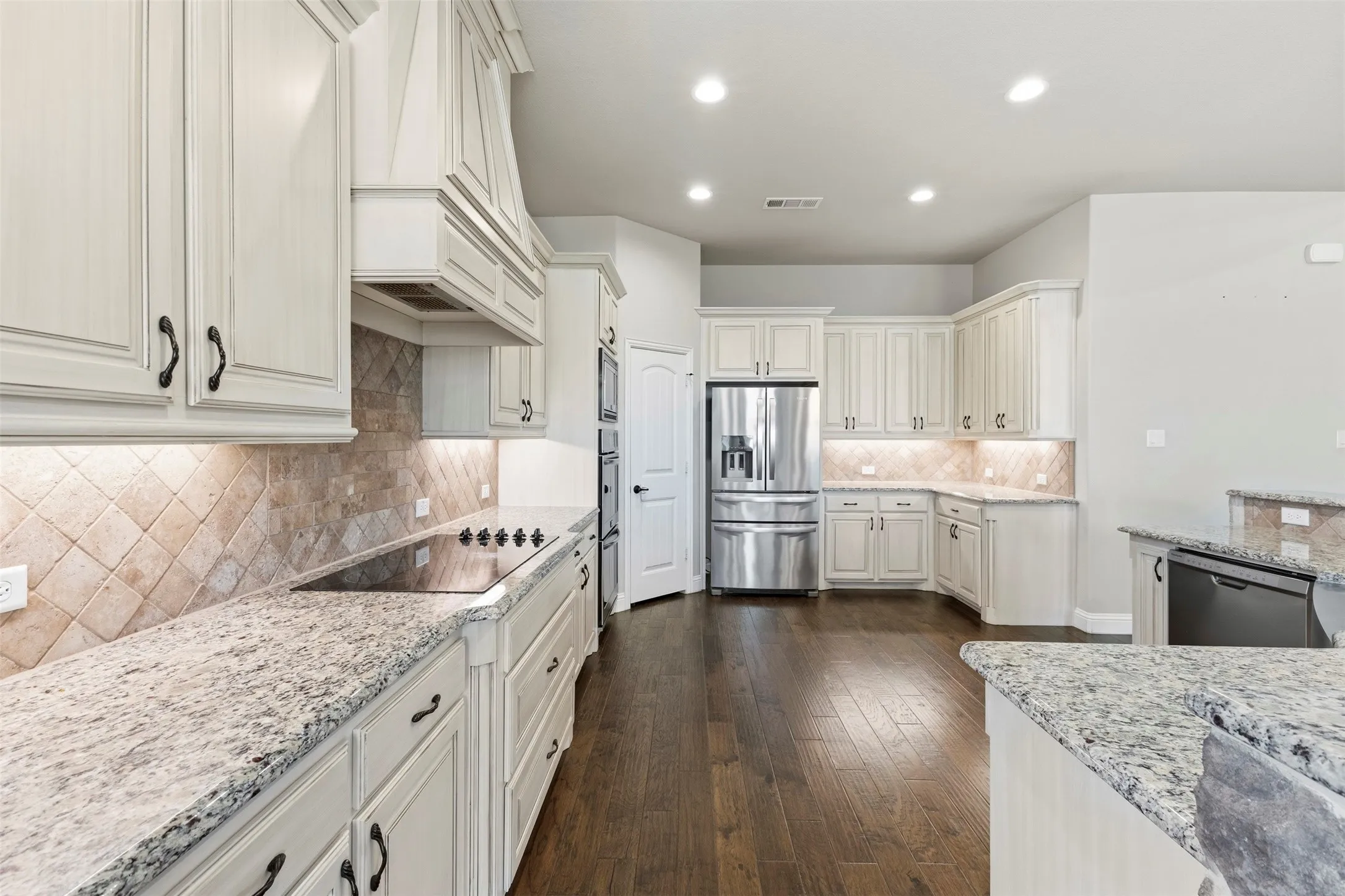 Chef's kitchen featuring white cabinetry with brushed nickel hardware, granite countertops, diagonal tile backsplash, stainless steel refrigerator and range, recessed lighting, and dark wood-finish flooring throughout the open layout