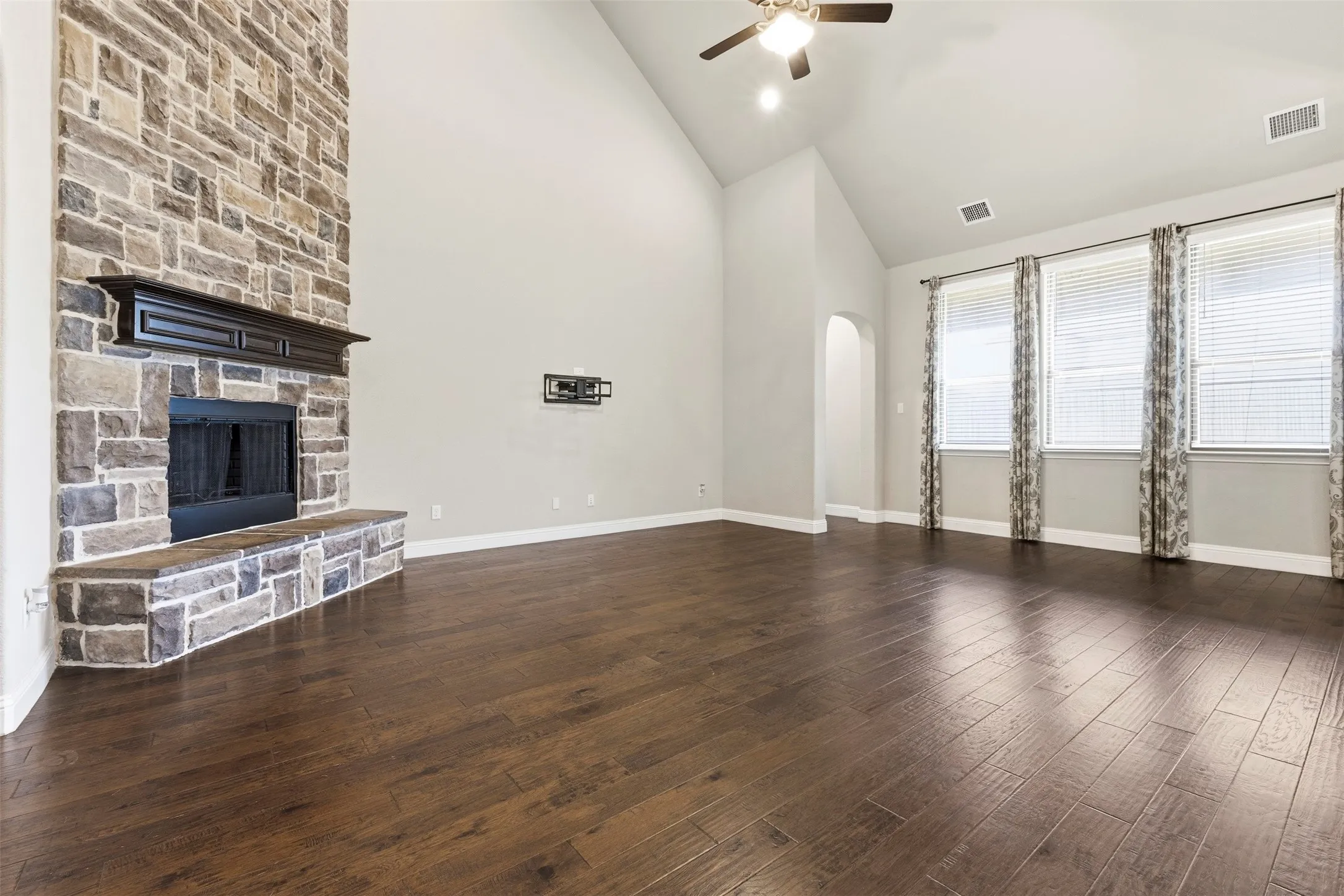 Spacious living area featuring a stone fireplace with a dark wood mantel, high vaulted ceilings, wood-finish flooring, and multiple windows