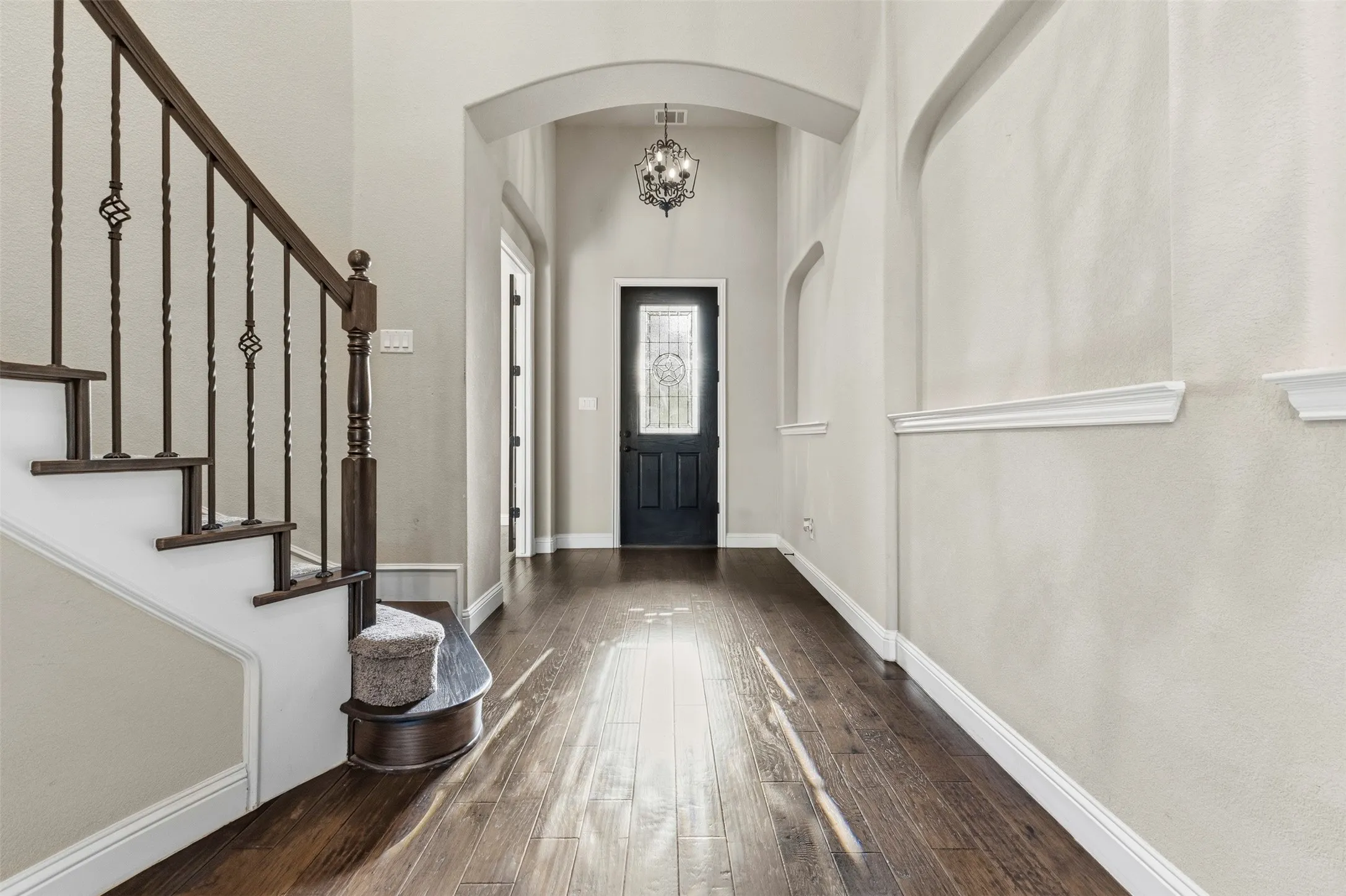 Welcoming foyer featuring a curved archway, dark wood-finish flooring, white shiplap wainscoting, a staircase with ornamental iron railings and wood newels, and a black entry door with sidelights topped by a decorative pendant fixture