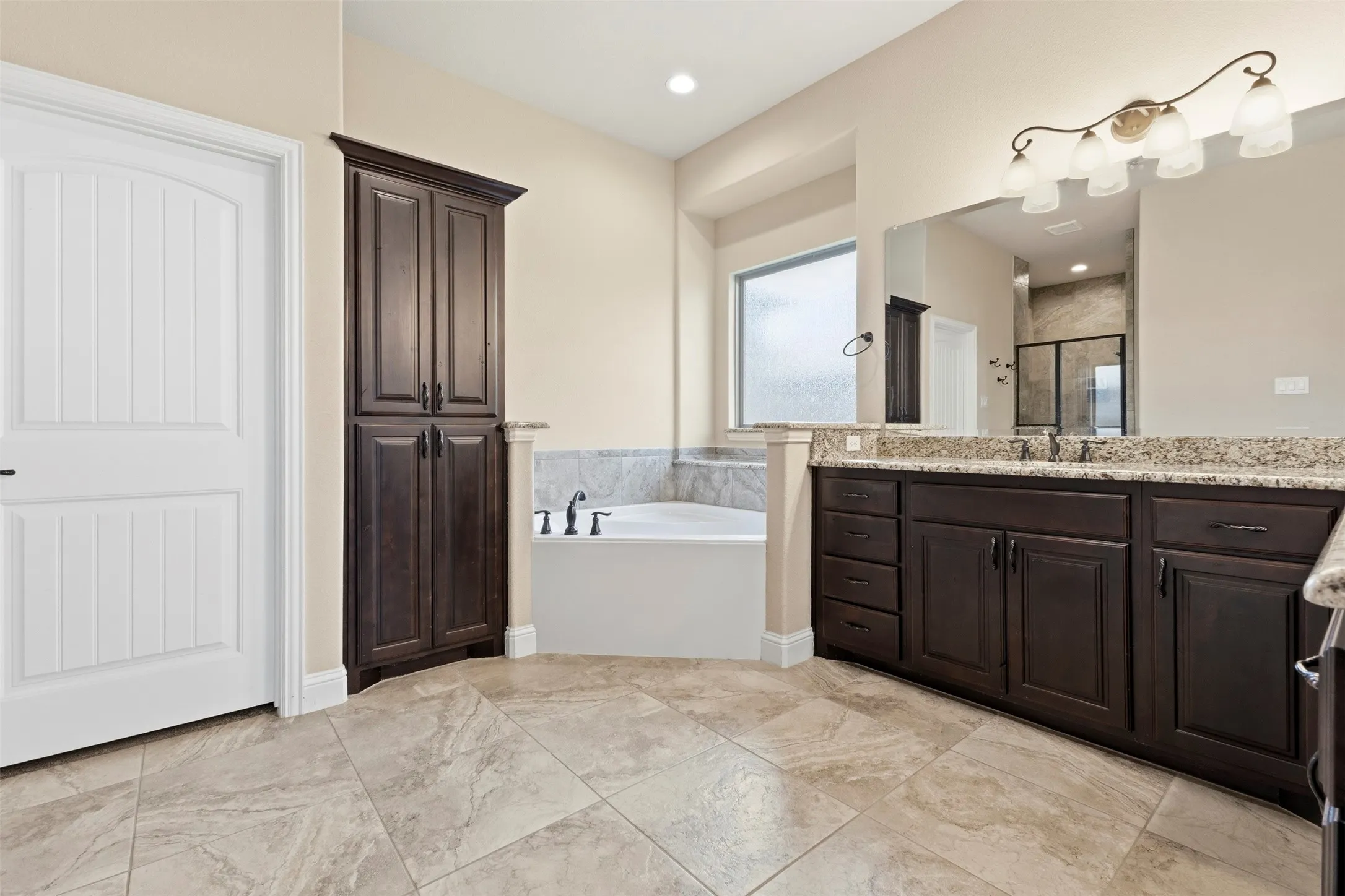 Spa-inspired master bath featuring dual vanities with granite countertops, dark cabinetry with ample storage, a soaking tub with tile surround, and large framed mirror with integrated lighting