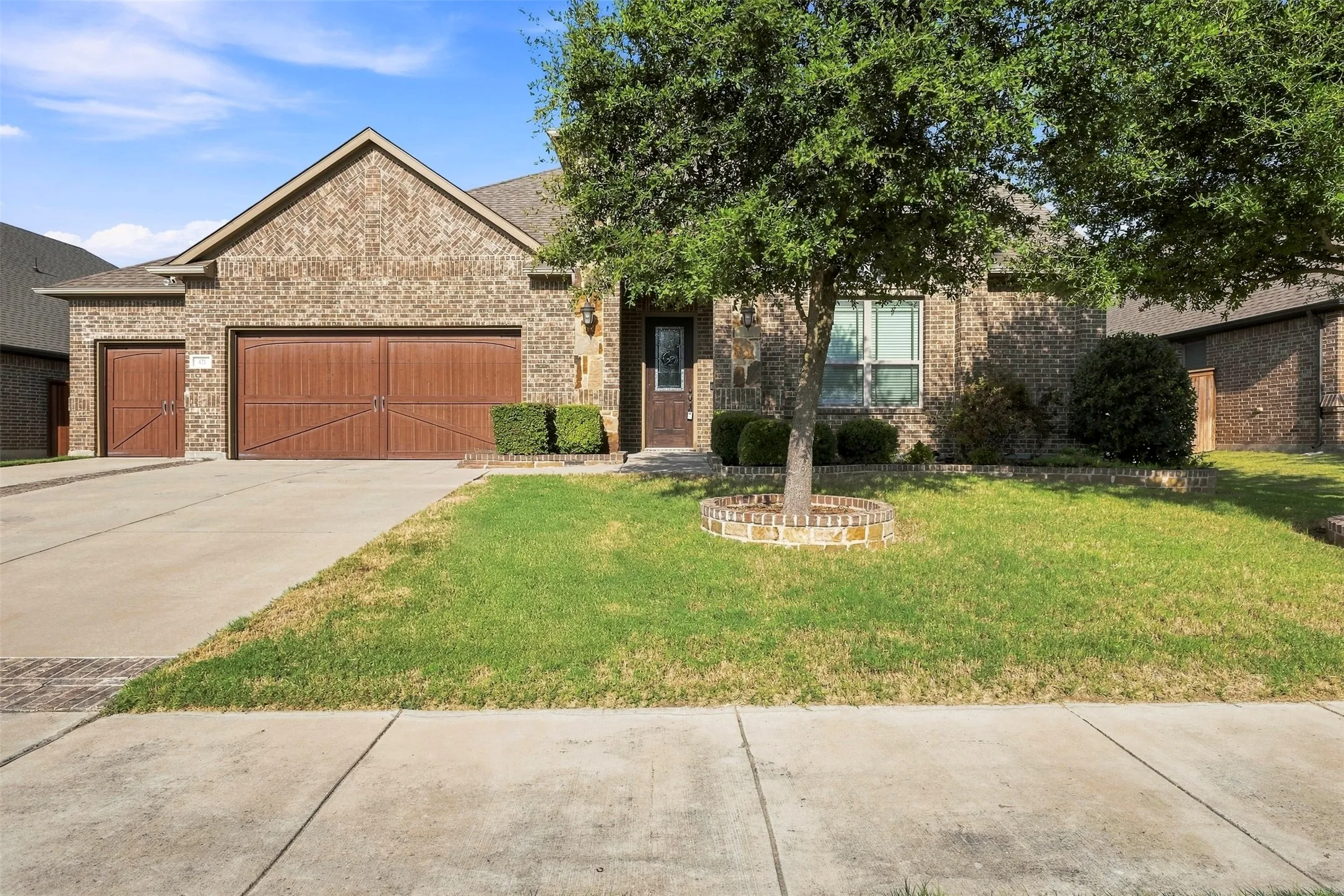 Brick exterior with a gabled roof, two wood-finish garage doors, concrete driveway, and front lawn with mature trees
