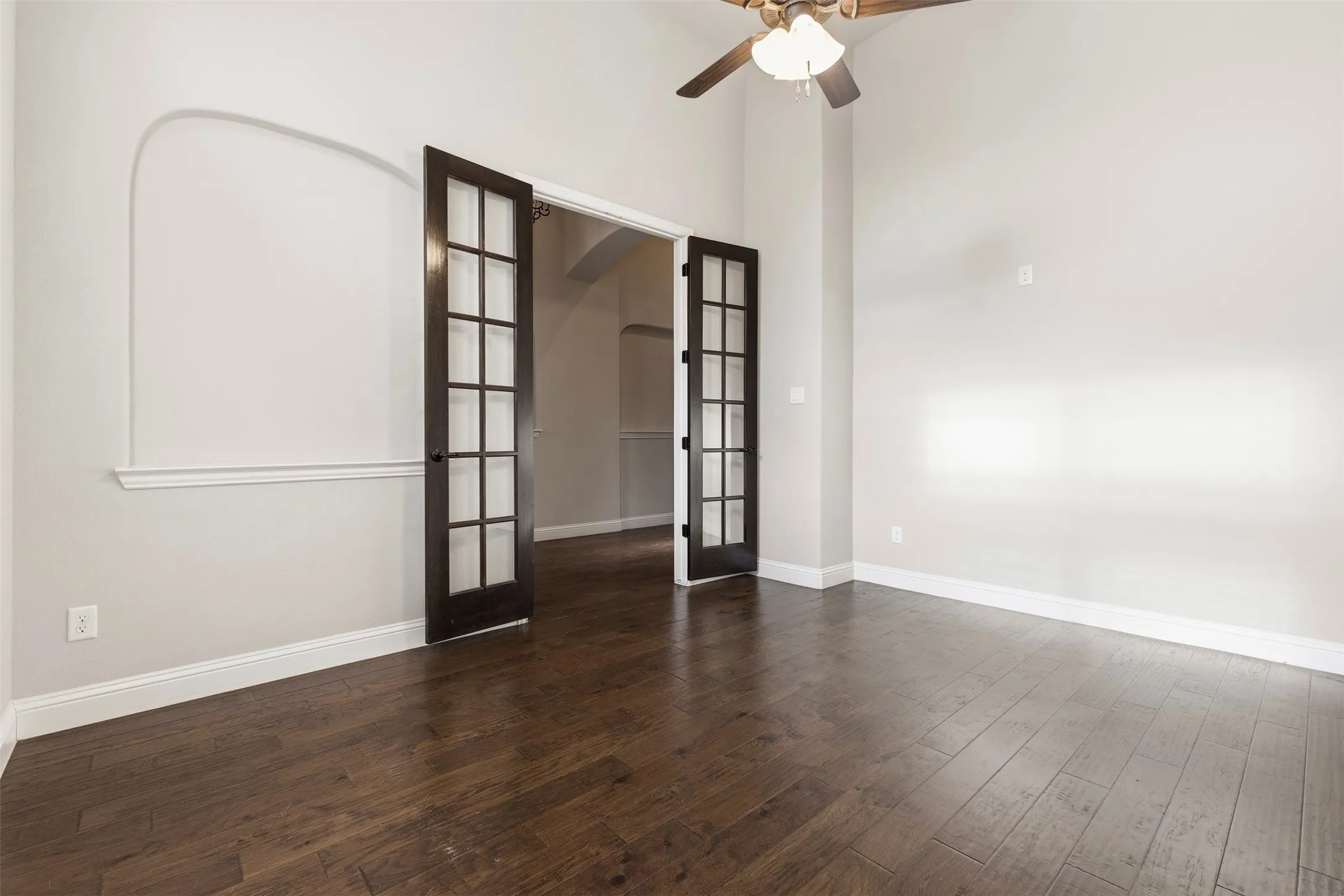Spacious room showcasing rich wood-finish flooring, ceiling fan with integrated lighting, and French doors with decorative glass panes opening to adjacent spaces