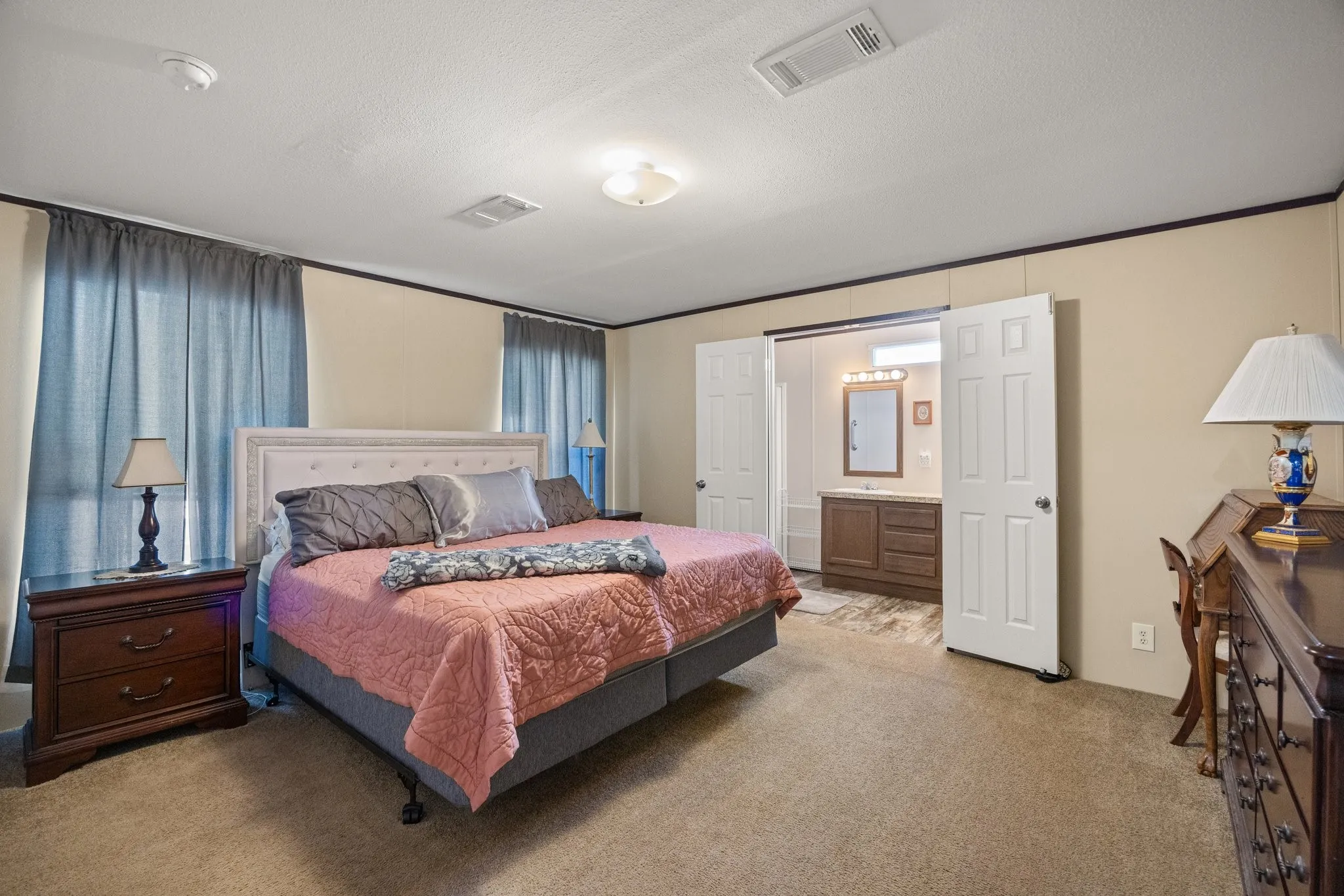Carpeted bedroom featuring an en-suite bathroom with a wood-finish vanity, a wall-mounted mirror, and overhead lighting