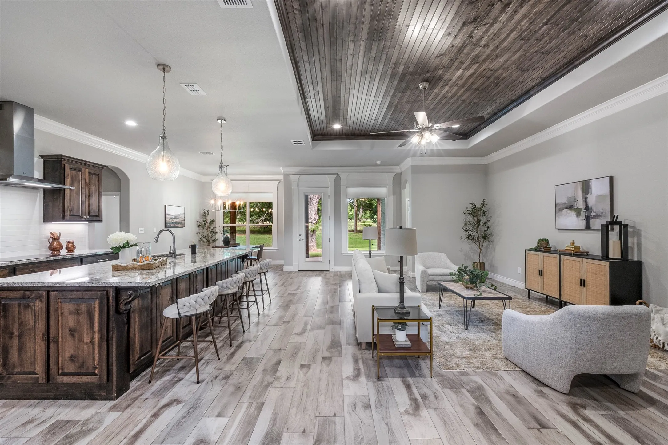 Open-concept kitchen and living room with large granite island, custom stained cabinetry, exposed wood ceiling and wood-look tile flooring.