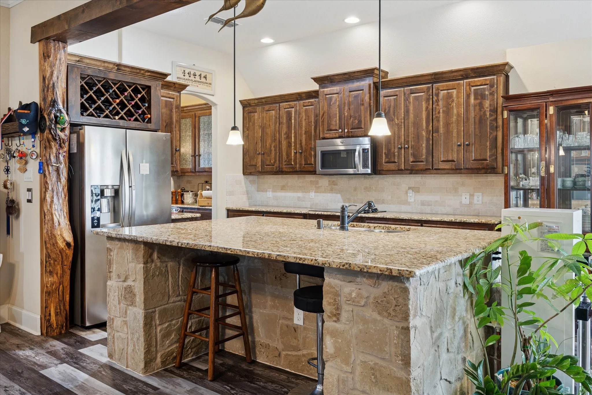 Kitchen featuring an expansive stone-clad island with a sink, pendant lighting, wood-finish cabinetry, a tile backsplash, and stainless steel appliances