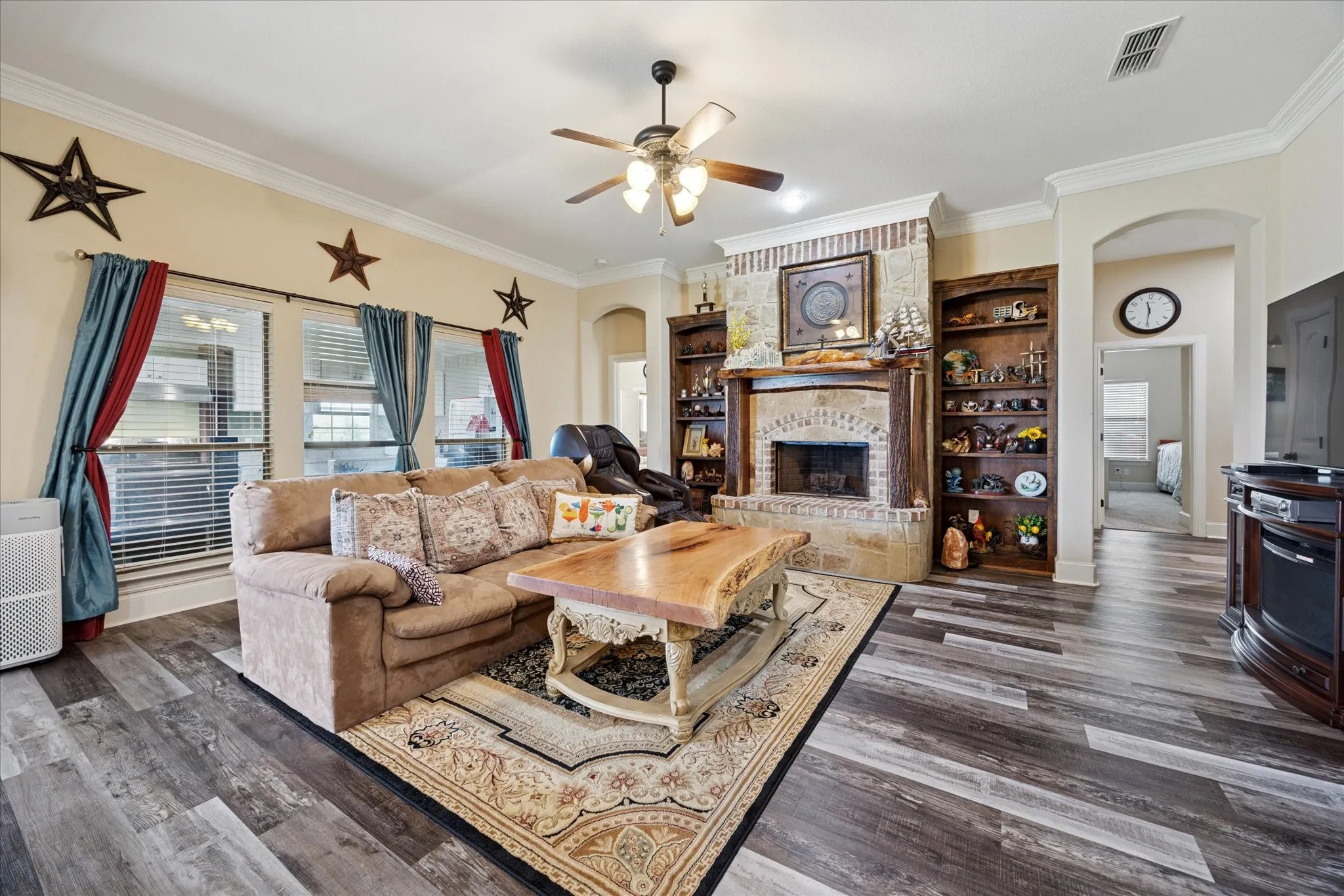 Spacious living area featuring wood-finish flooring, an arched brick fireplace with built-in shelving, decorative crown molding, and multiple windows with blinds