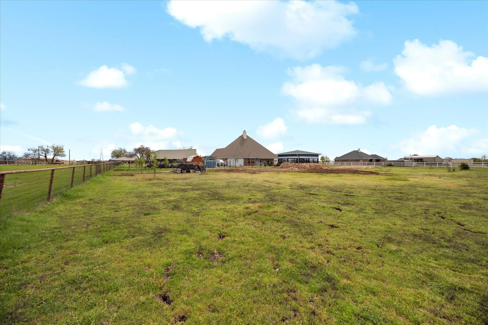 Expansive green acreage with perimeter fencing, featuring several single-story structures with varied rooflines in the background