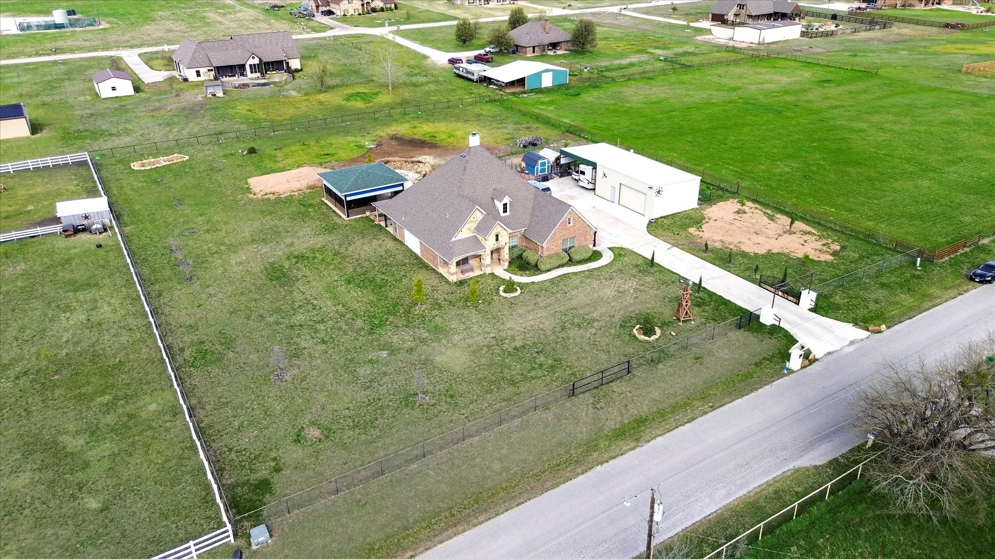 Spacious property featuring a brick-clad residence with a multi-gabled roofline, an expansive green lawn, an attached covered patio, a detached garage structure, and a concrete driveway