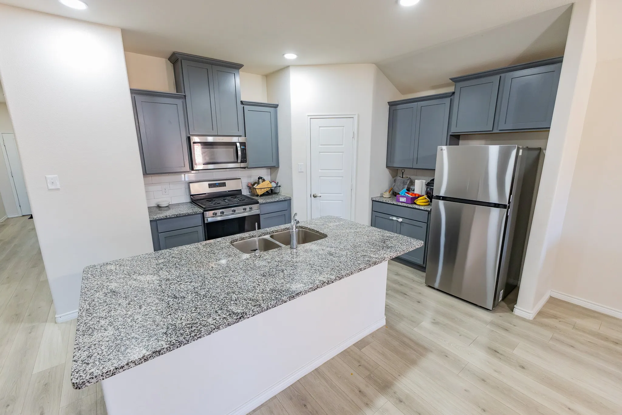 Kitchen featuring an island with a double basin stainless steel sink, granite countertops, gray cabinetry, stainless steel appliances, and wood-finish flooring