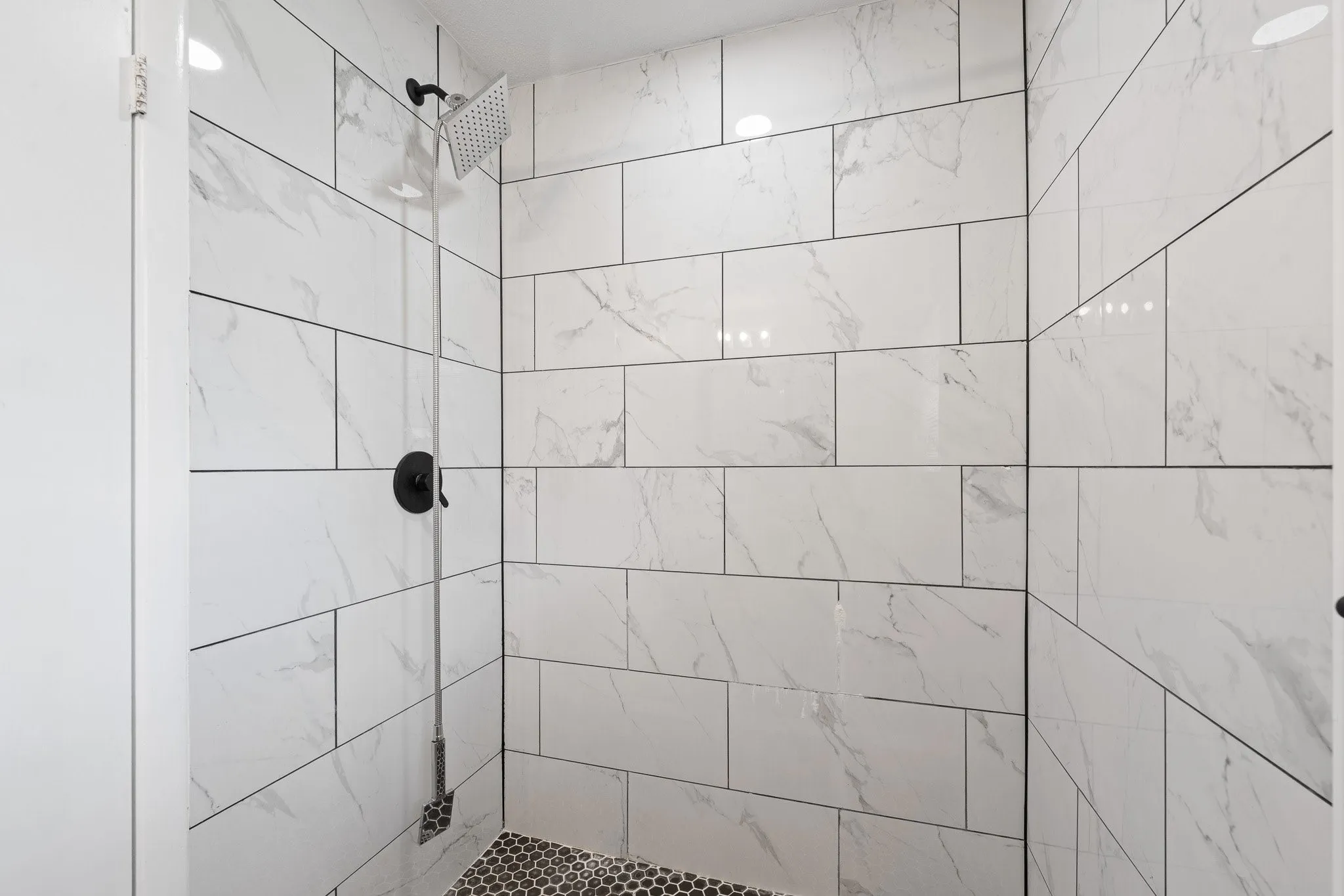 large walk in Shower featuring white subway tile with subtle grey veining, a matte black rain shower head, matte black shower controls, and a black penny tile floor