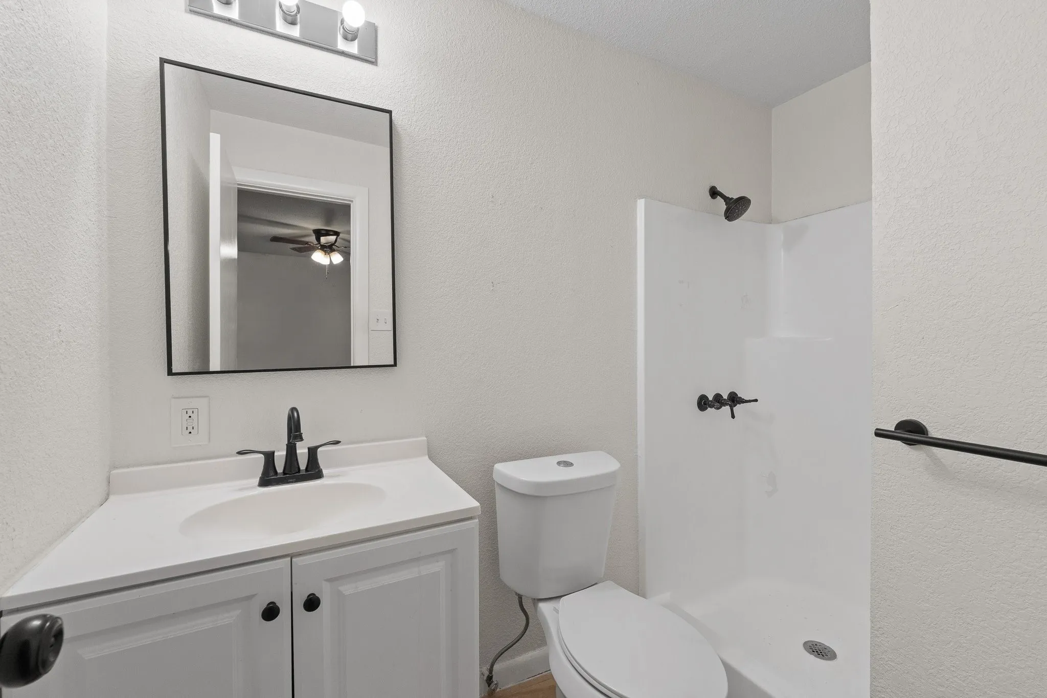 Bathroom featuring a white vanity with integrated sink, black fixtures, rectangular mirror, and a white shower stall with a black shower head and controls