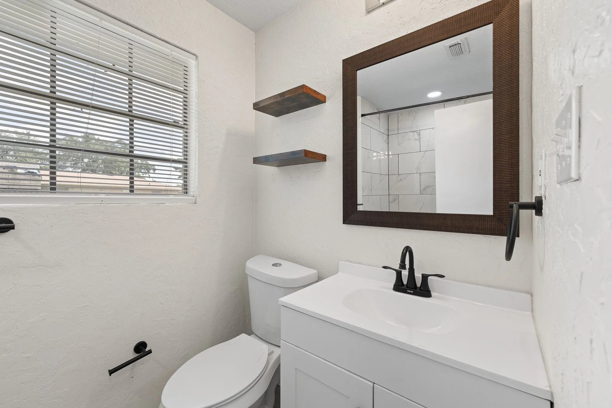 Bathroom featuring a white vanity with a dark bronze faucet, a framed mirror, and two wood-finish floating shelves