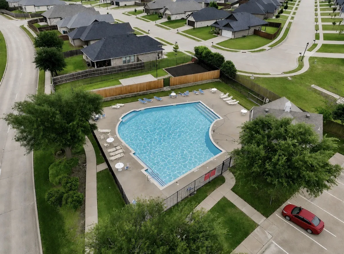 Expansive swimming pool with concrete decking, surrounded by a perimeter fence and mature trees