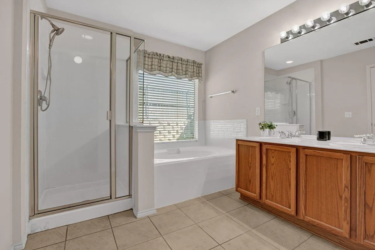 Bathroom featuring a glass-enclosed shower, a built-in soaking tub beneath a window, a double vanity with wood cabinetry, a large wall-mounted mirror, and tile flooring
