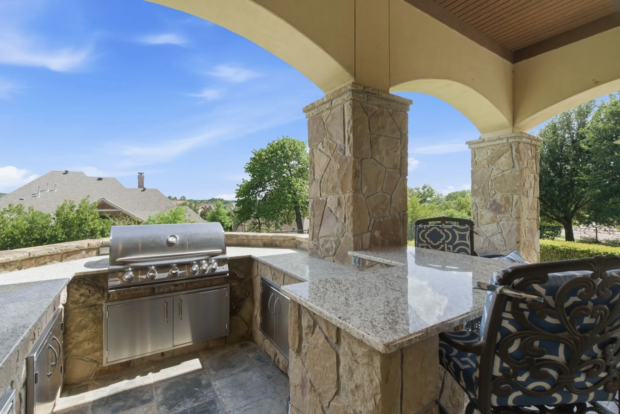 Stone-clad outdoor kitchen featuring a built-in stainless steel grill, granite countertops, and integrated cabinetry under an arched patio cover with architectural columns