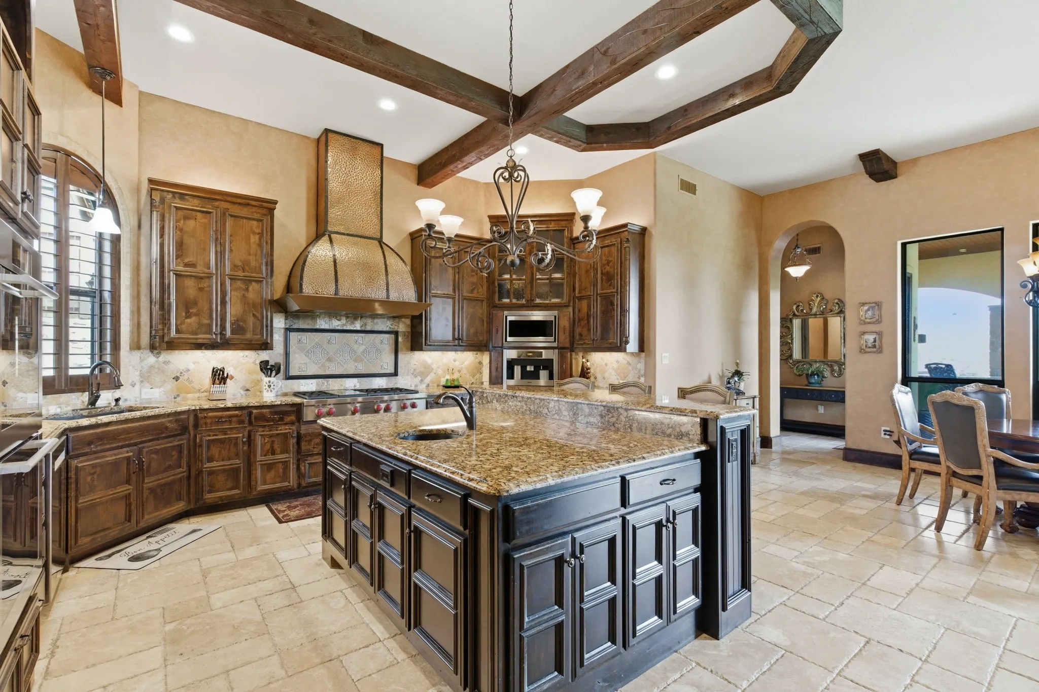 Spacious kitchen featuring an expansive center island with a granite countertop, custom wood cabinetry, a large decorative range hood, stone tile flooring, and exposed wood beam ceiling accents