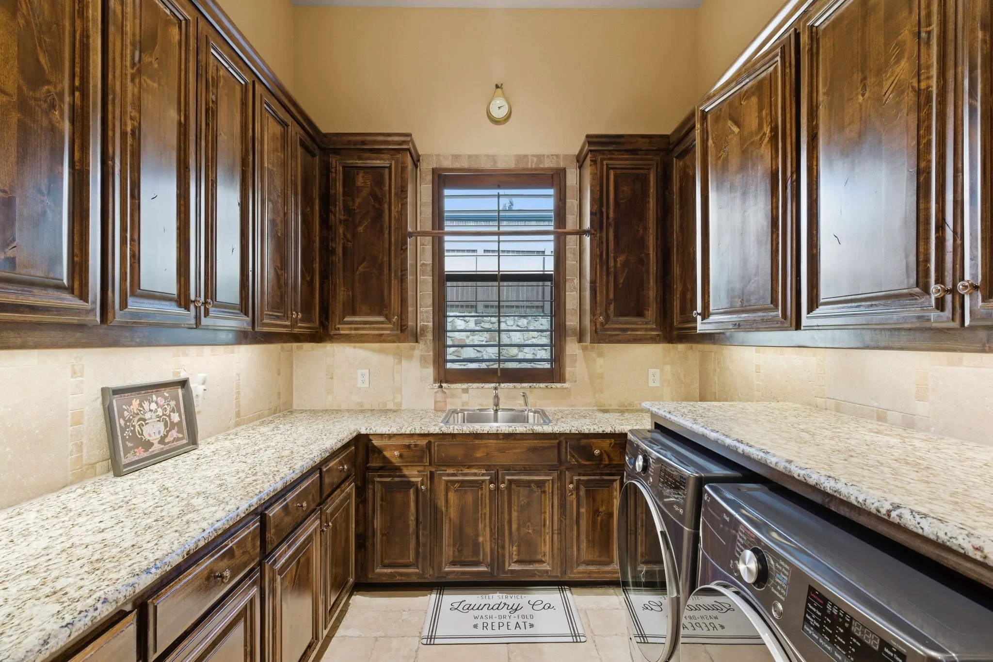 Utility room featuring extensive wood cabinetry, light-toned granite-finish countertops, a built-in sink, tiled flooring, and a window providing natural light