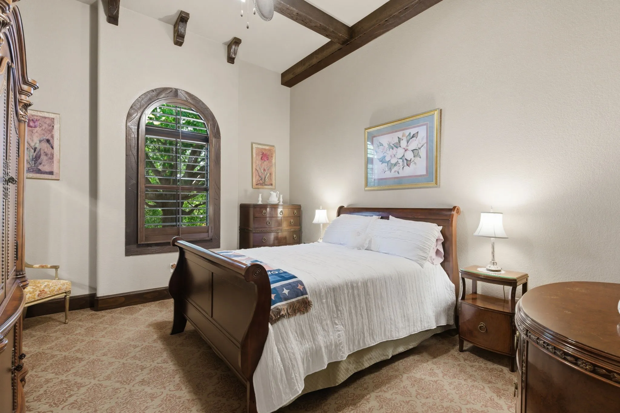 Downstairs Guest Suite Room featuring an arched window with wooden plantation shutters, exposed ceiling beams, decorative corbels, and patterned carpet flooring