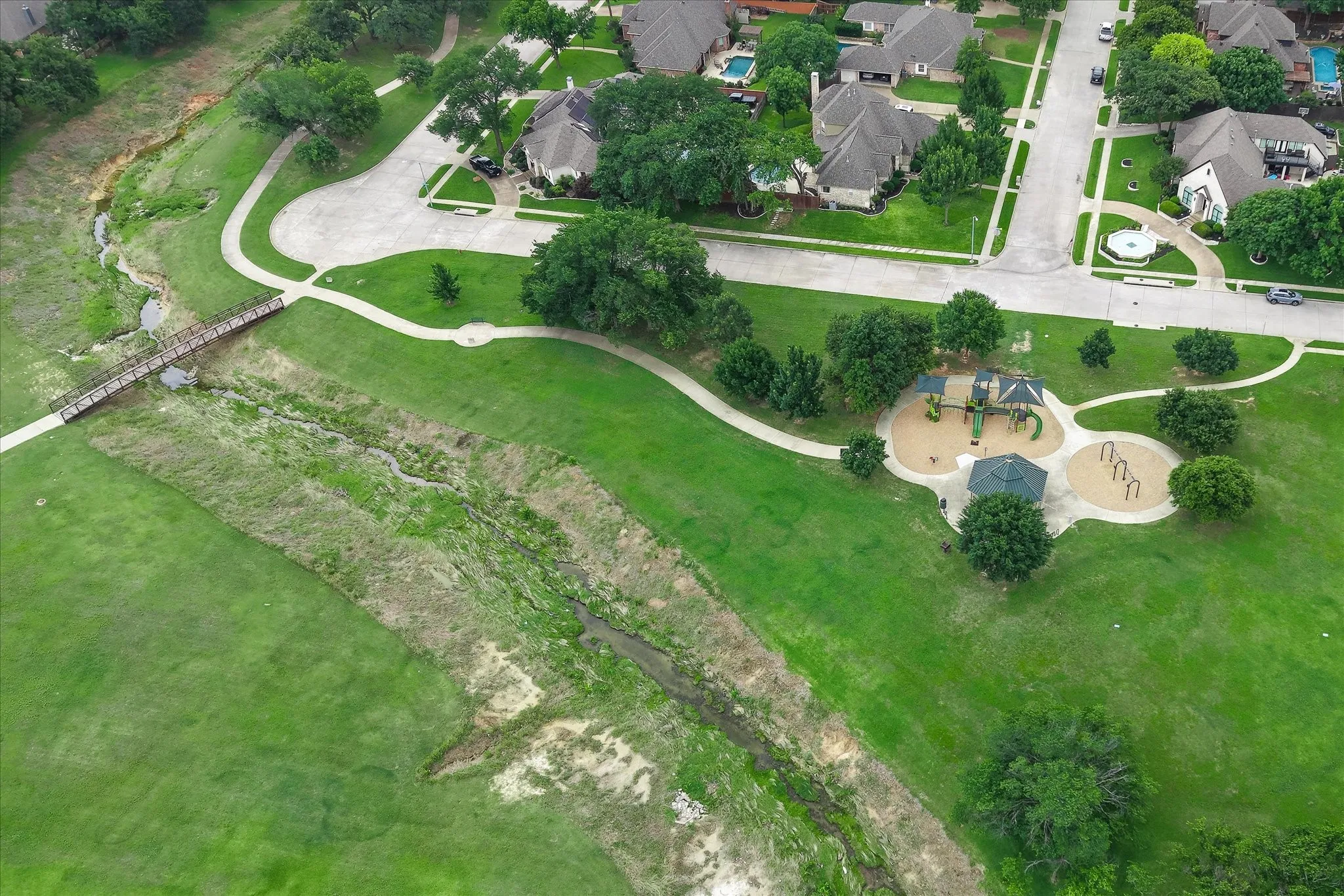 Expansive green space featuring a playground with multiple structures, a shaded gazebo, winding paved pathways, a pedestrian bridge over a natural creek, and mature trees