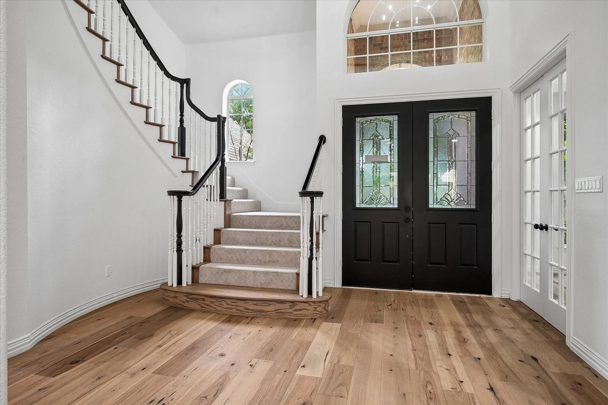 Grand entryway featuring a double staircase, wood-finish flooring, black double entry doors with leaded glass, an arched transom window, and a white paneled French door