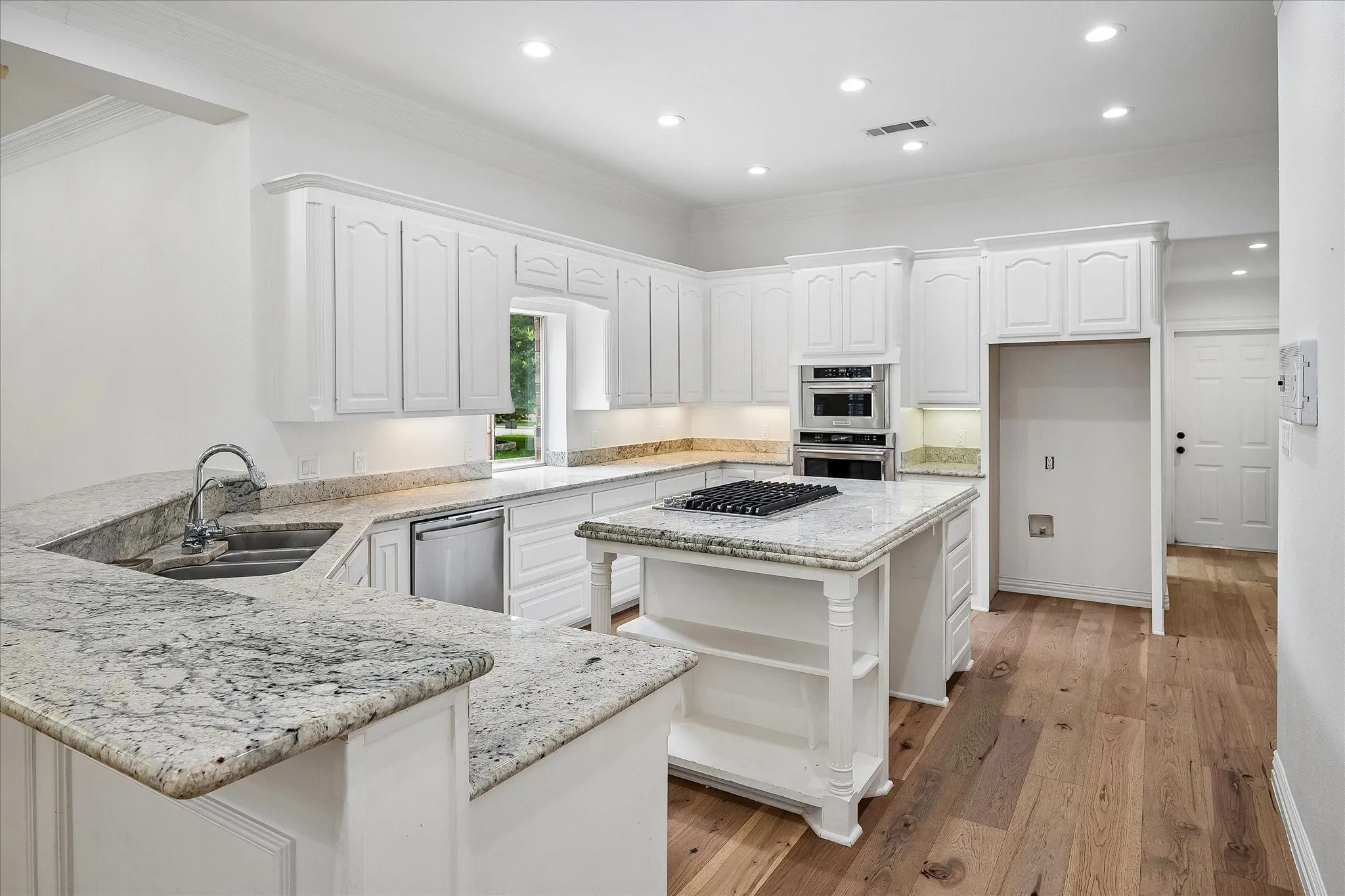 Spacious kitchen featuring white cabinetry, light-toned stone countertops, a central island with an integrated cooktop, built-in double ovens, and wood-finish flooring