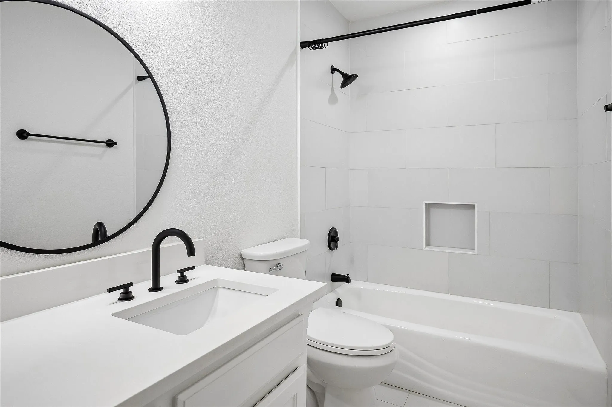 Bathroom featuring a white vanity with an integrated sink and matte black fixtures, a circular mirror, a white bathtub with subway tile surround, and a recessed shower niche