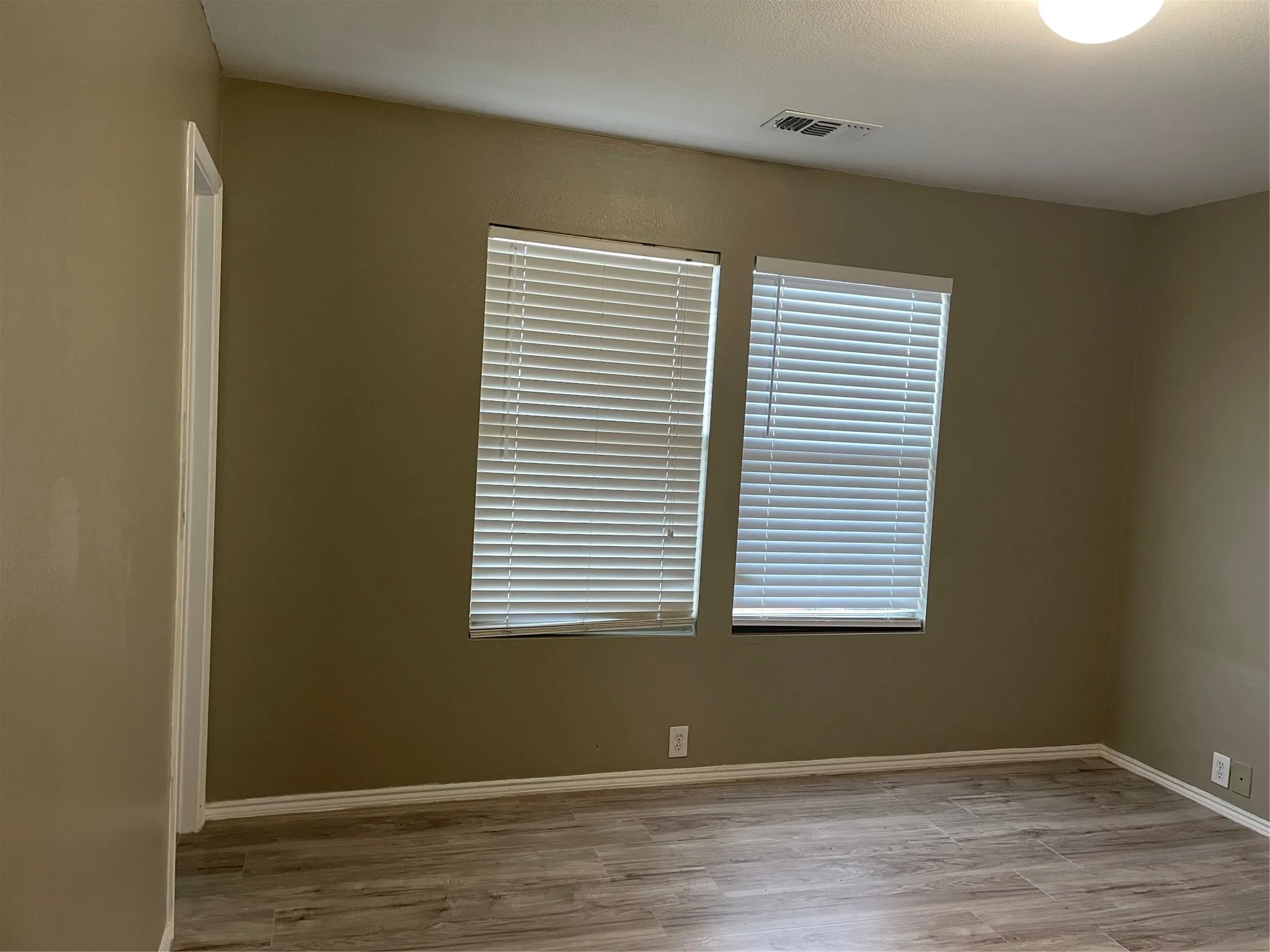 Interior space featuring wood-finish flooring, two windows with blinds, a white ceiling light fixture, and neutral painted walls