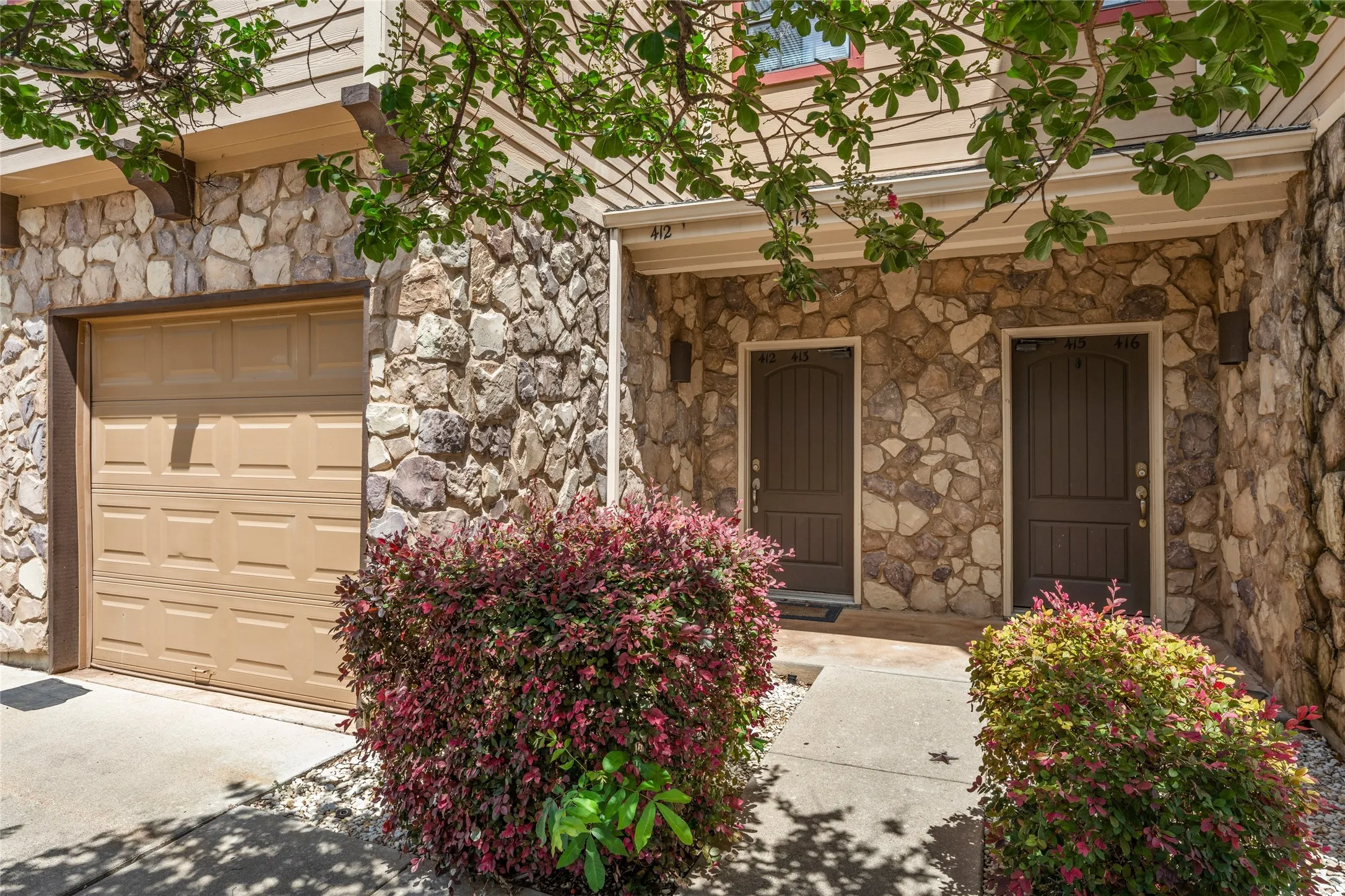 Stone facade exterior featuring a tan garage door, two arched entry doors, and established landscaping