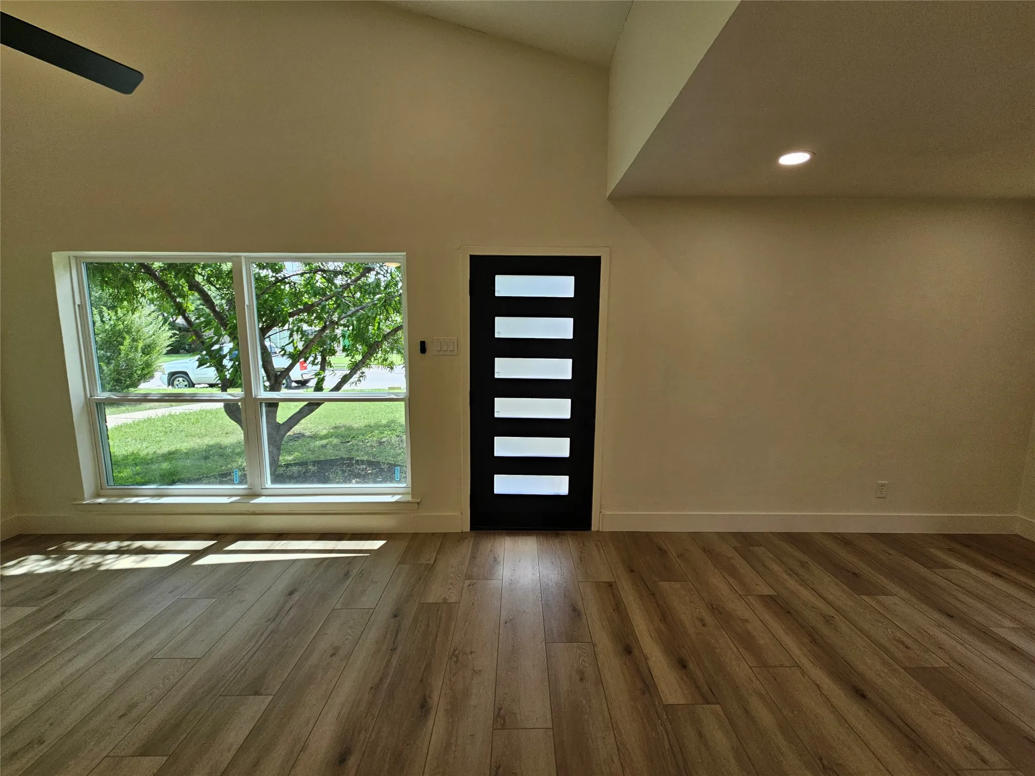 Spacious living area featuring high vaulted ceilings, wood-finish flooring, a contemporary entry door with frosted glass inserts, recessed lighting, and a large picture window