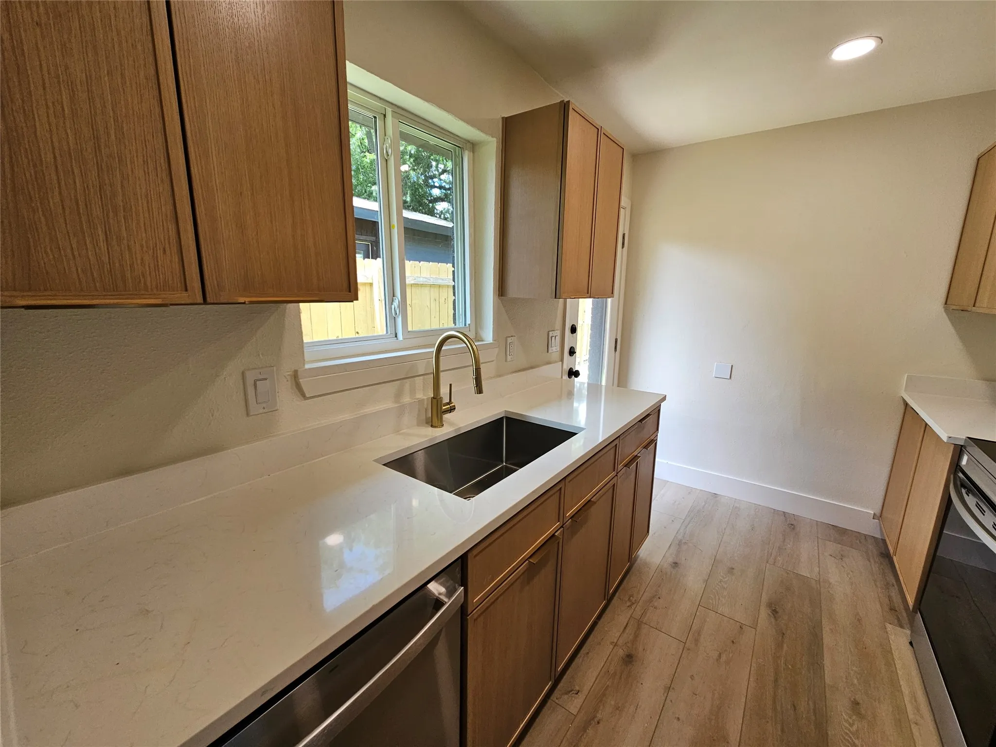 Modern kitchen featuring wood-finish cabinetry, light-toned countertops, a stainless steel undermount sink, and a gold-tone gooseneck faucet
