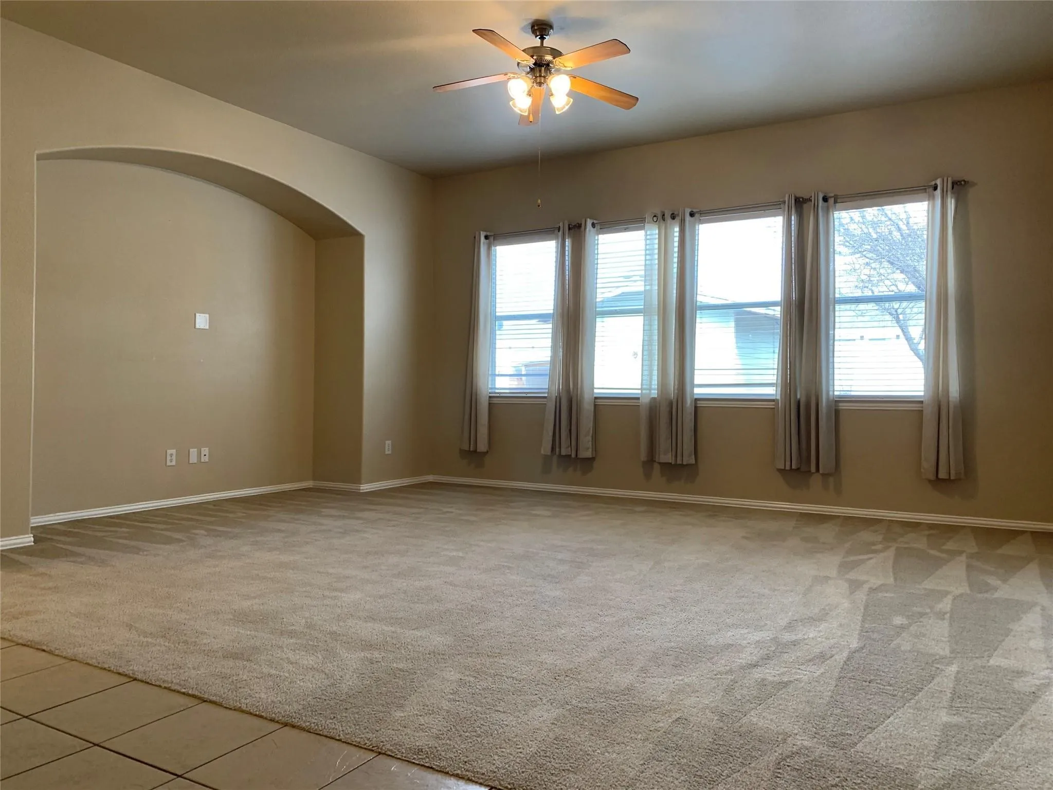 Spacious room featuring neutral carpeting, a ceiling fan with light fixture, and a wall of four windows with blinds and drapes