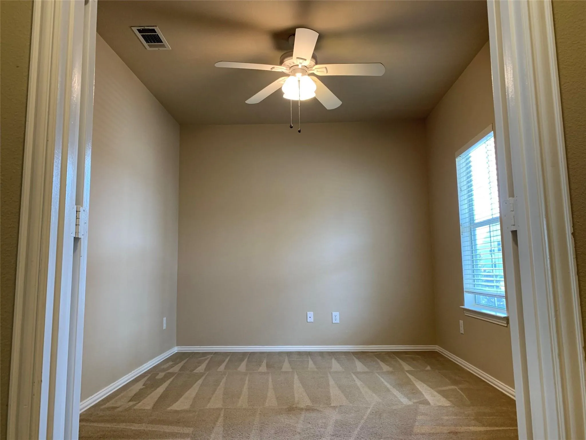 Carpeted interior room featuring neutral wall tones, a ceiling fan with integrated lighting, and a single window with blinds