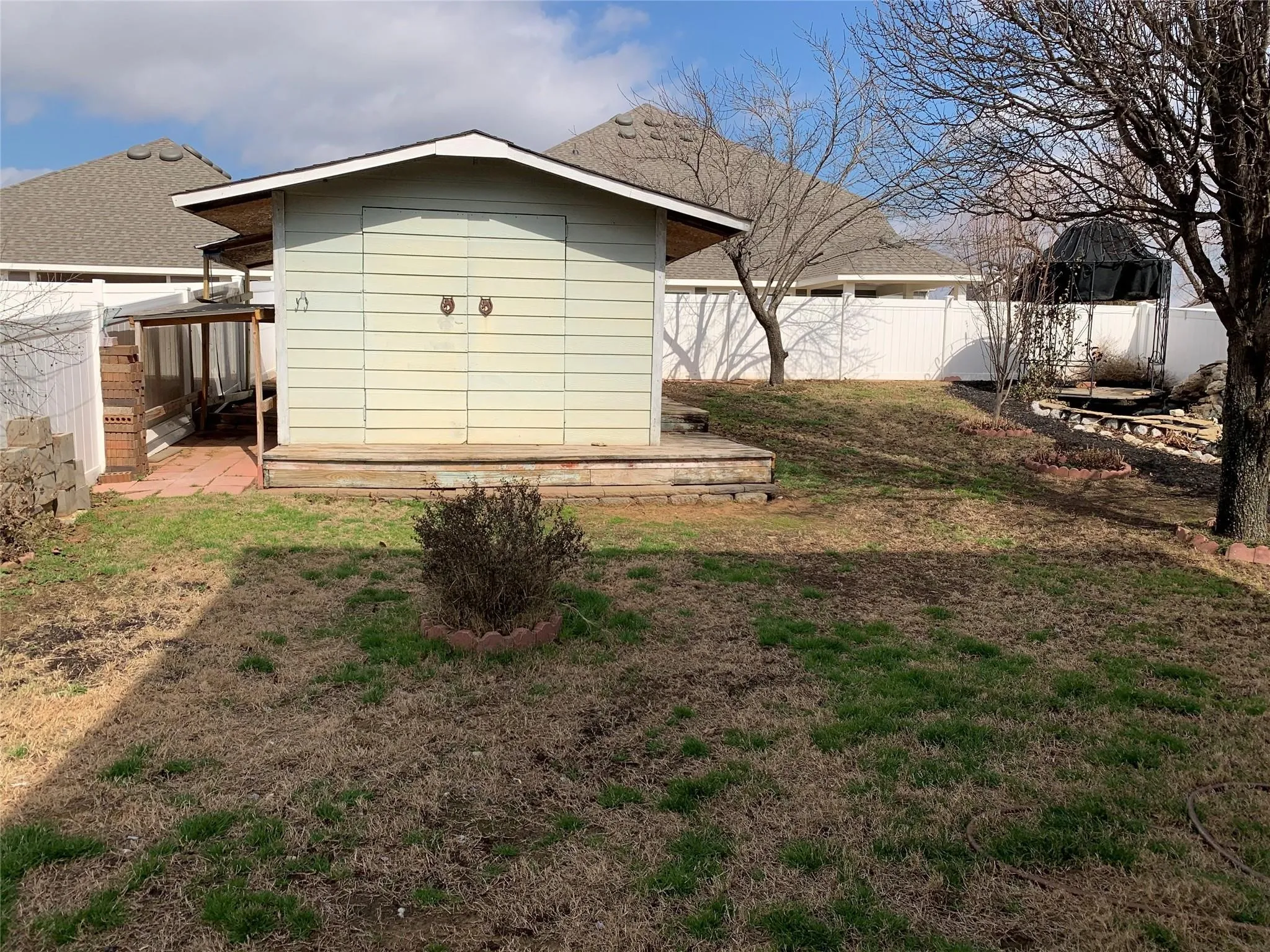 Backyard featuring a light green storage shed with double doors, a white privacy fence, a covered patio area with reddish-brown paving, and a sloped terrain
