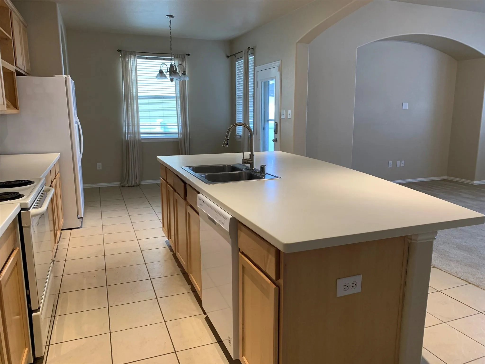 Kitchen featuring an island with double basin sink, light-toned countertops, blonde wood cabinetry, and white appliances