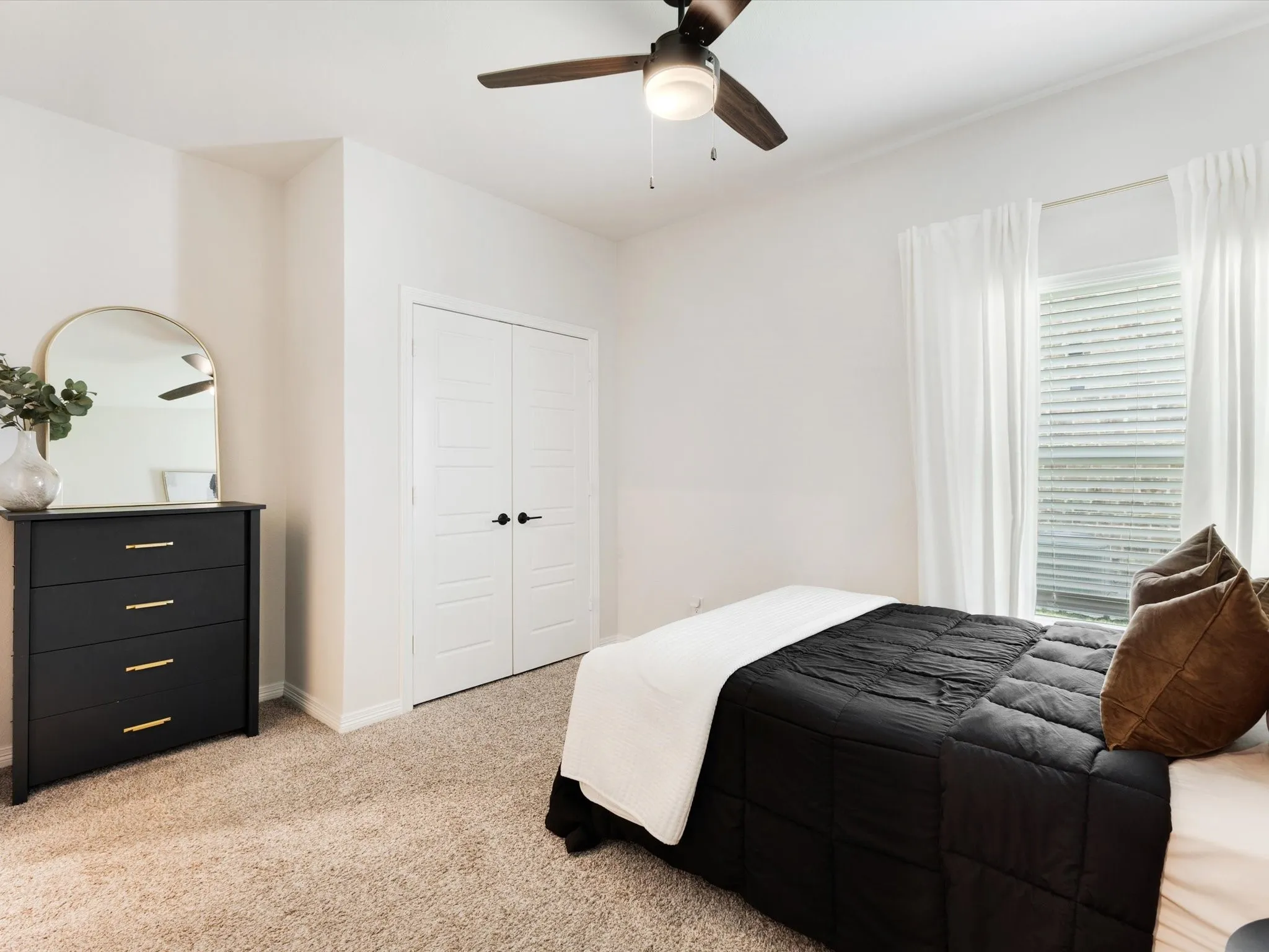 Neutral-toned bedroom featuring a ceiling fan with light fixture, window with blinds and sheer curtains, carpet flooring, and white double closet doors