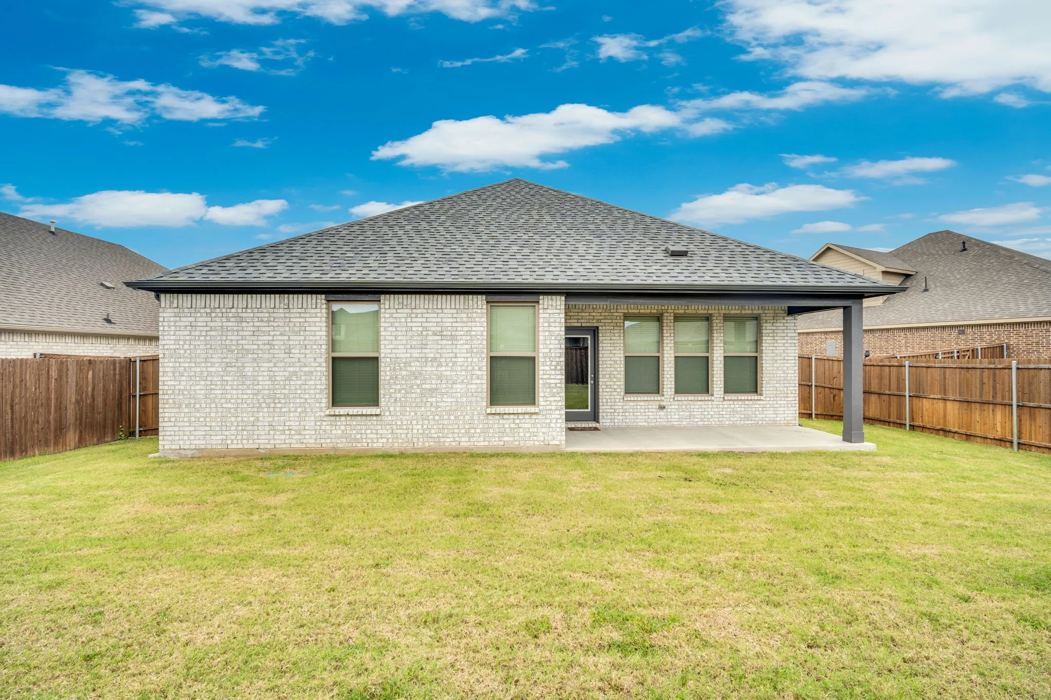 Expansive grassy backyard featuring a light-colored brick exterior, covered patio, dark grey trim, and a privacy fence