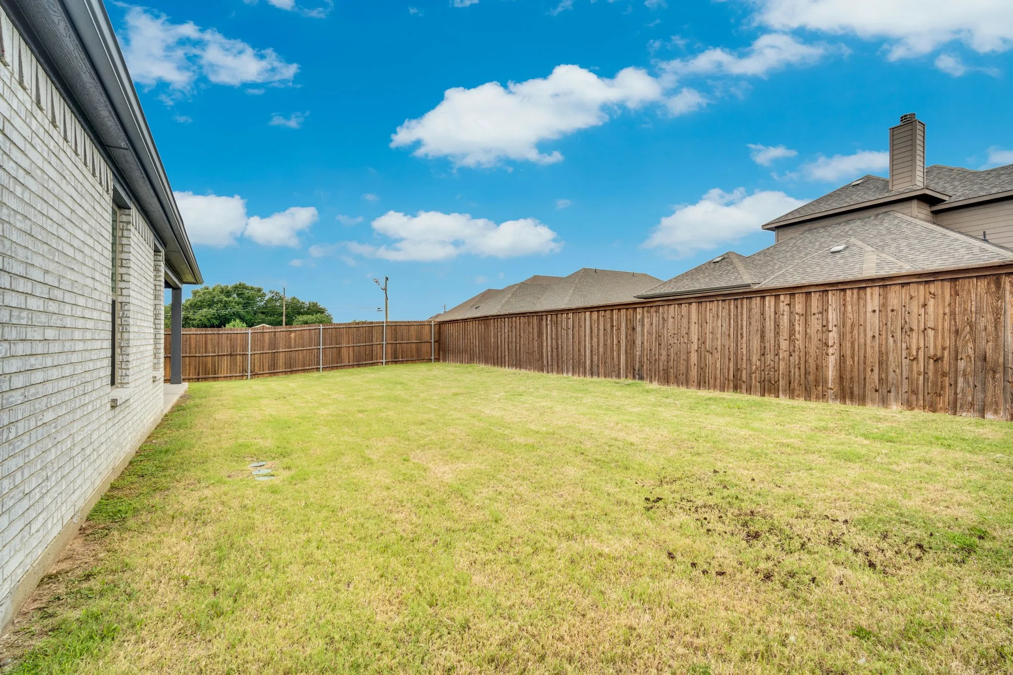 Expansive grassy backyard featuring a light-colored brick exterior, covered patio, dark grey trim, and a privacy fence