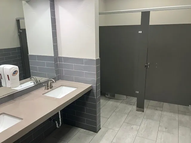Spacious restroom featuring dual integrated sinks with brushed nickel fixtures, a neutral-toned countertop, and gray subway tile wainscoting