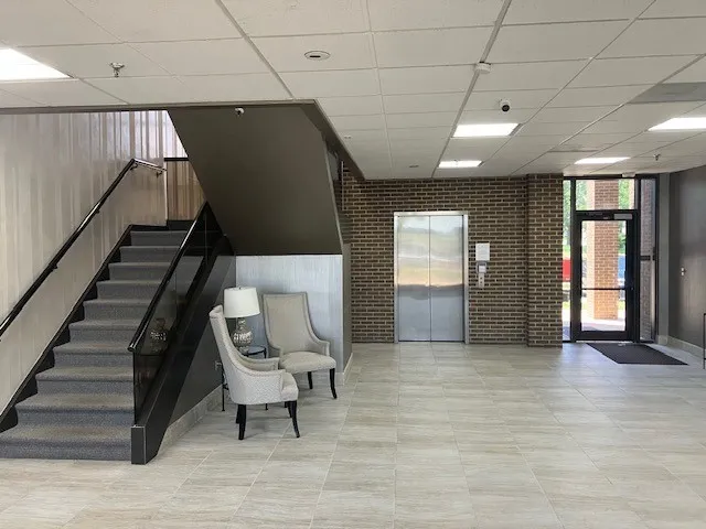 Lobby entrance featuring a staircase with black railing, a stainless steel elevator, brick accent wall, and light-toned tile flooring
